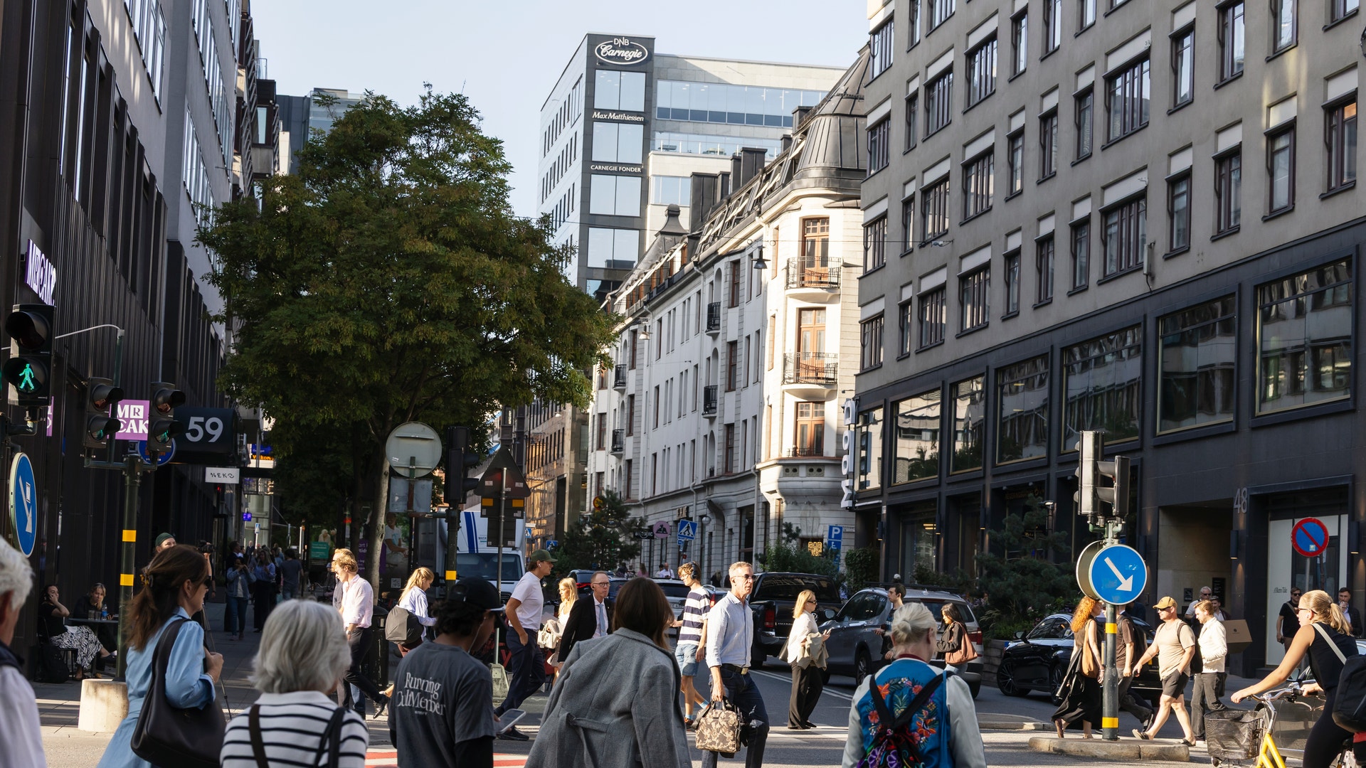 En livlig stadsgata med många människor som går omkring mellan klassiska och moderna byggnader. Människor i olika kläder går på en bred gata med cykelväg, omgivna av butiker och träd.