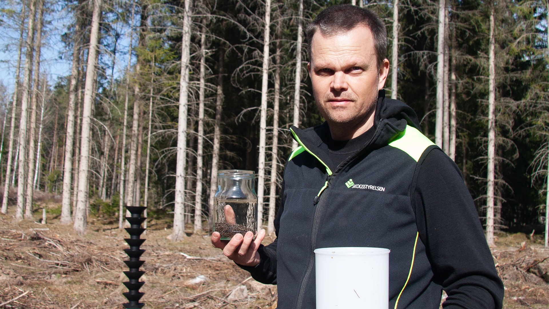 Man i skogen som håller upp en glaskruka. Han bär en svart jacka med vita och gula detaljer. Höga barrträd syns i bakgrunden.