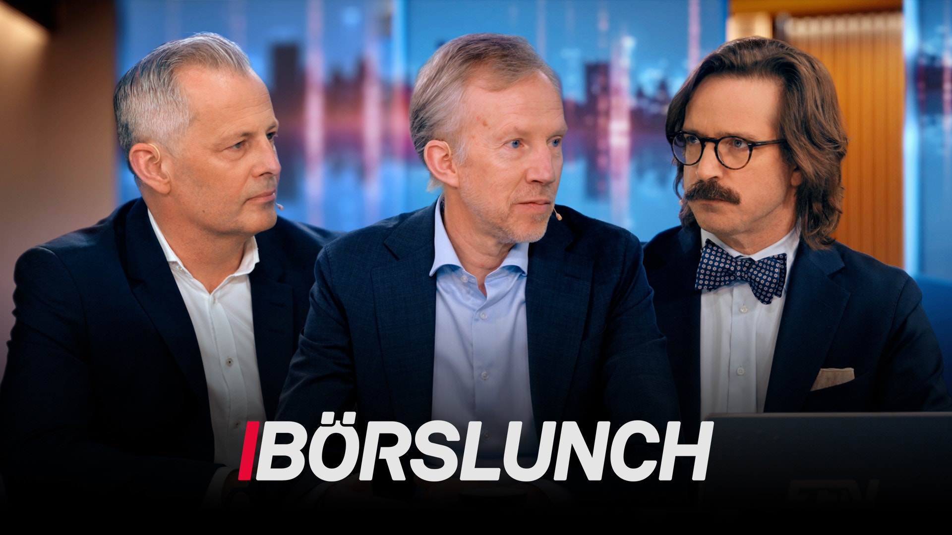 Tre män i affärskläder sitter tillsammans i en TV-studio med stadsvy i bakgrunden.