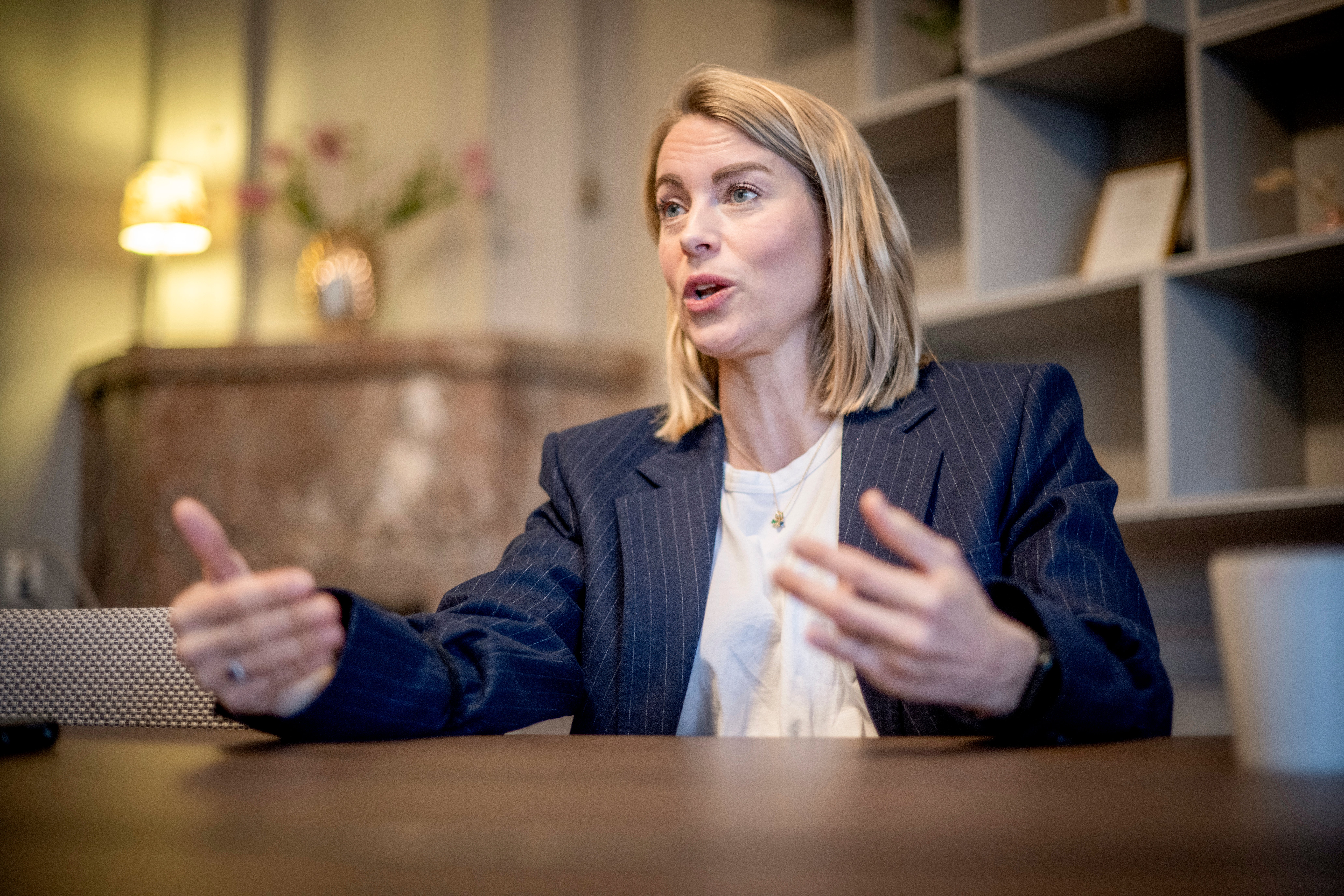 En kvinna i mörk kostym sitter vid ett skrivbord och gestikulerar medan hon talar.