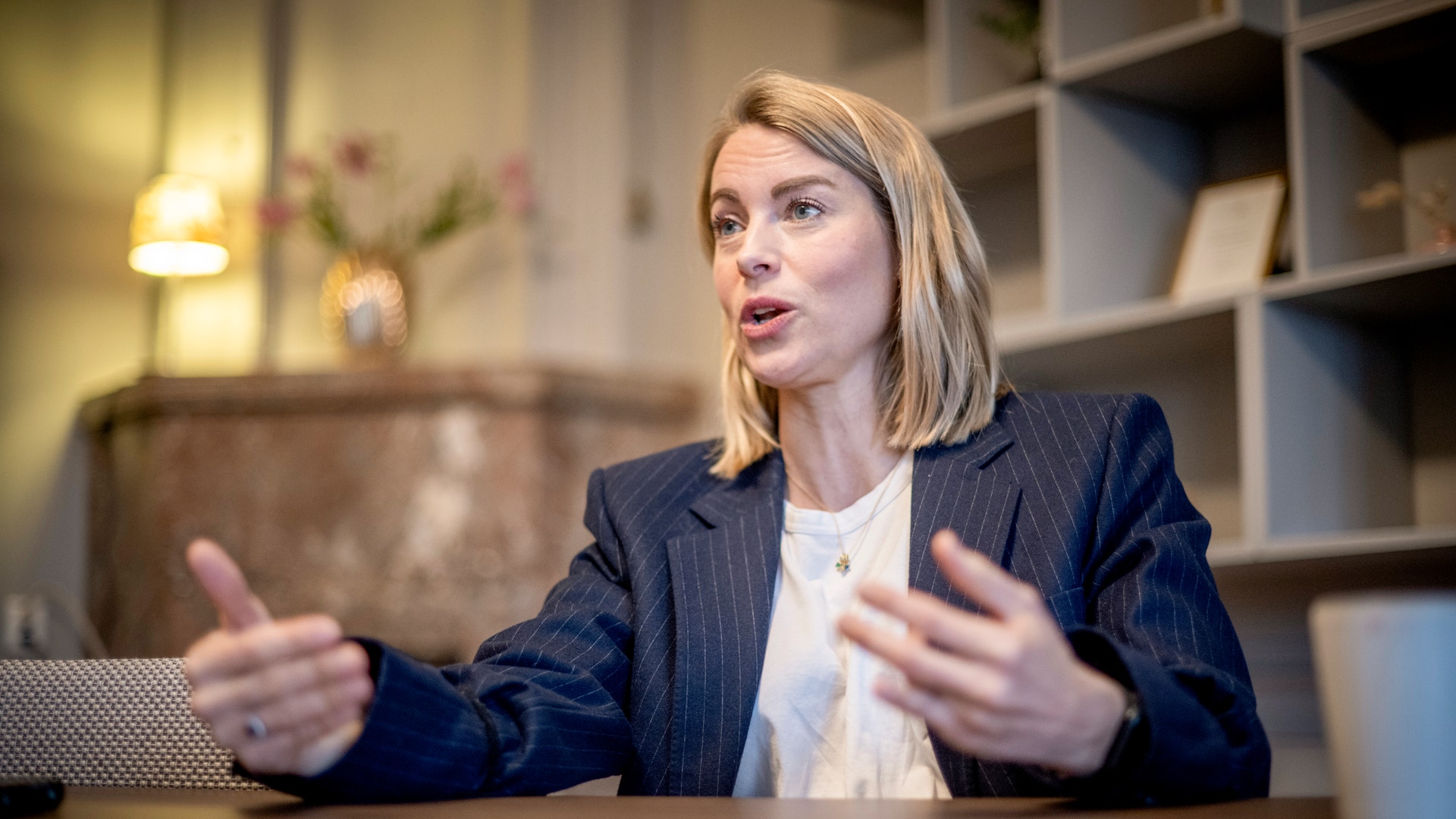 En kvinna i mörk kostym sitter vid ett skrivbord och gestikulerar medan hon talar.