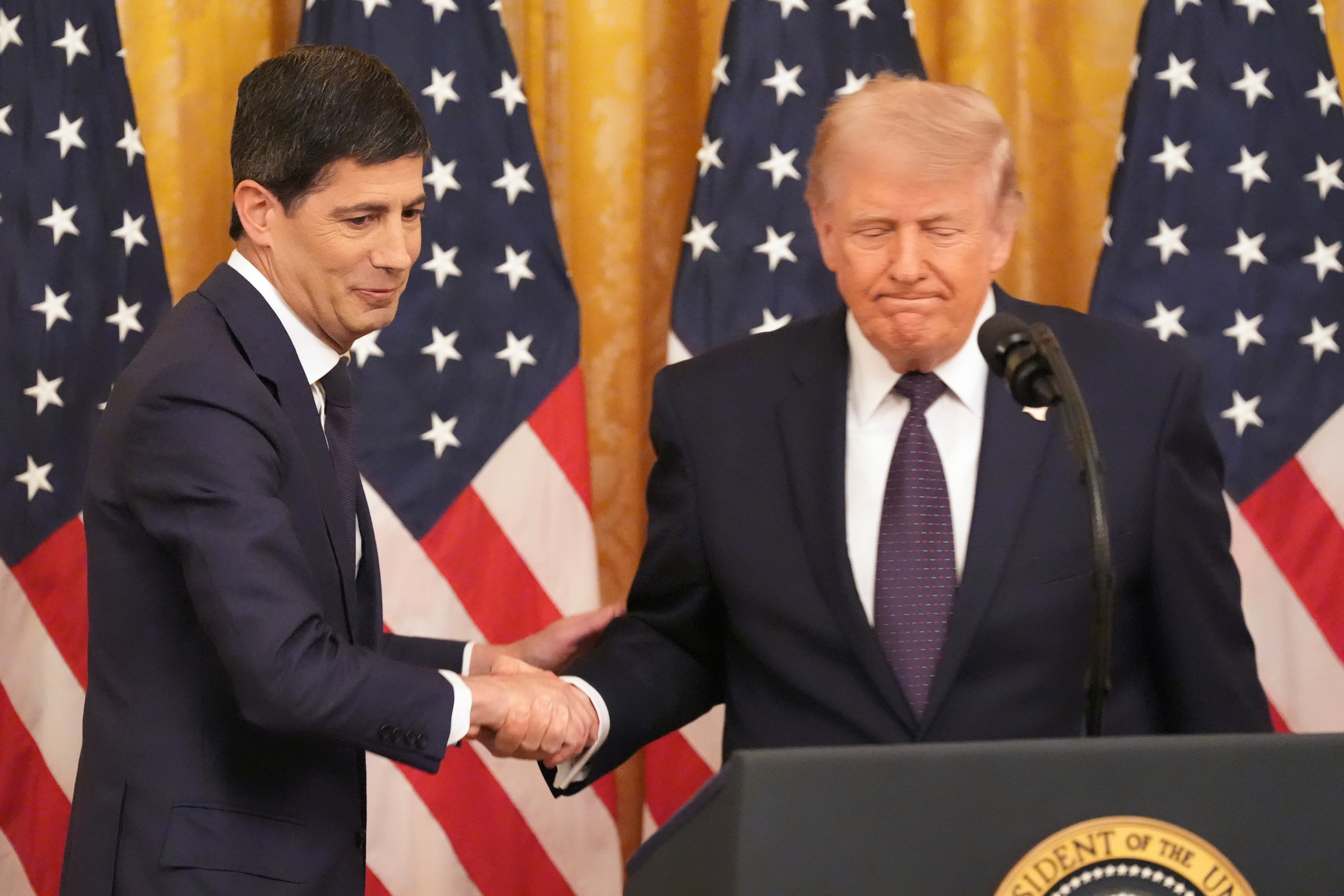 Två män i kostym skakar hand framför amerikanska flaggor vid ett officiellt möte.