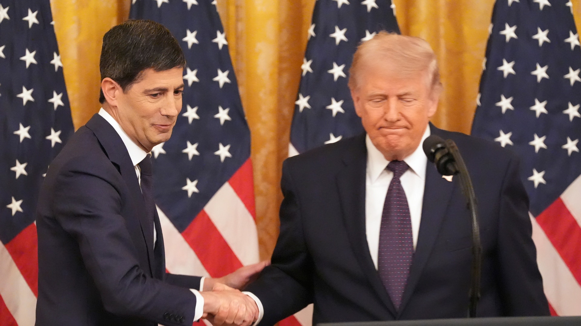 Två män i kostym skakar hand framför amerikanska flaggor vid ett officiellt möte.
