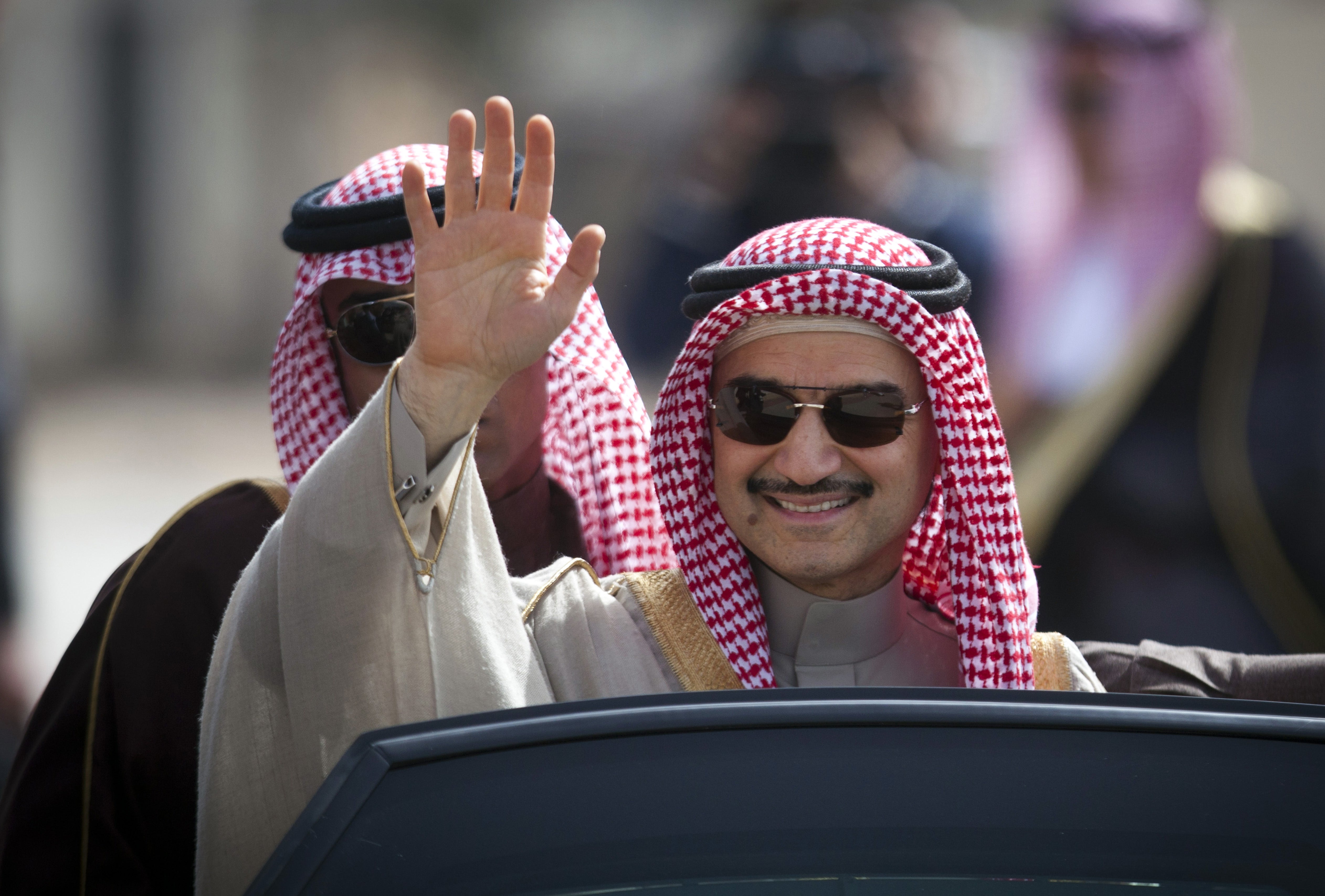 Två män i traditionell arabisk kläder sitter i en bil. Mannen i förgrunden vinkar och bär solglasögon.