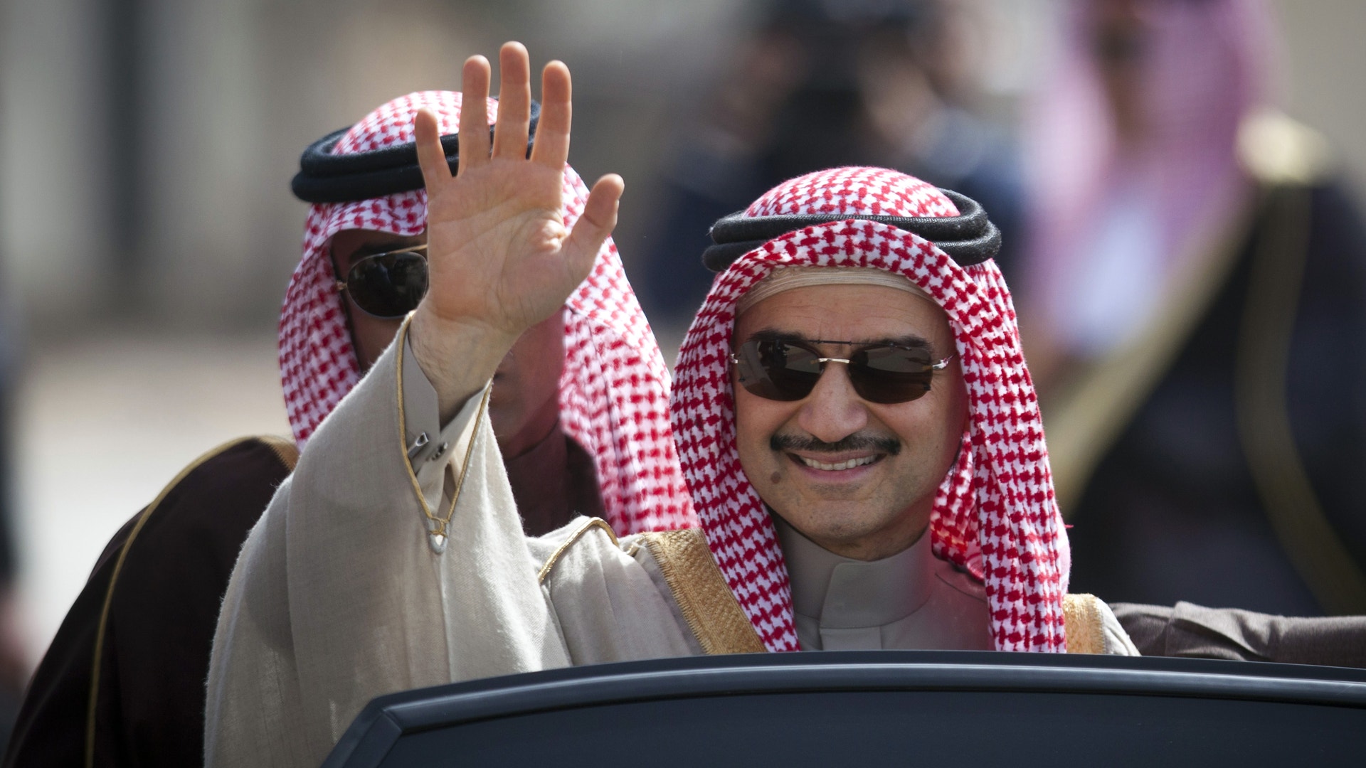 Två män i traditionell arabisk kläder sitter i en bil. Mannen i förgrunden vinkar och bär solglasögon.