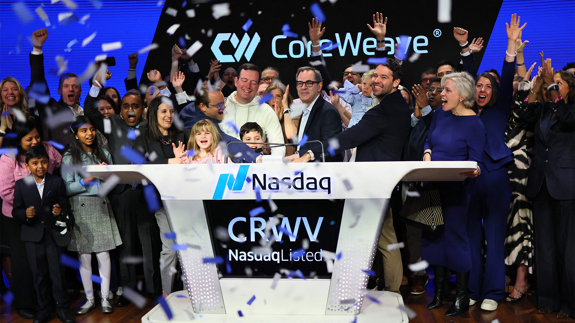 Jublande människor, bland dem barn, står vid en Nasdaq-podium under en börsnotering för CoreWeave. Konfetti faller i luften.