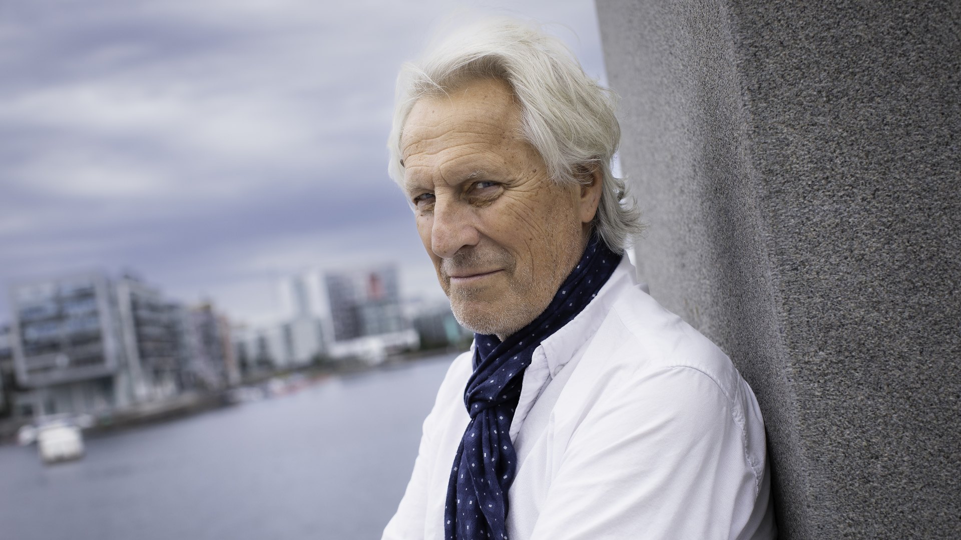 Porträtt av äldre man med vitt hår, vit skjorta och mörk slips framför en stadssiluett vid vatten.