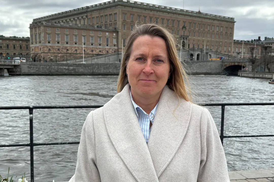 Porträtt av en kvinna i ljus kappa som står framför en vattenbild med historiska stenbyggnader i bakgrunden.