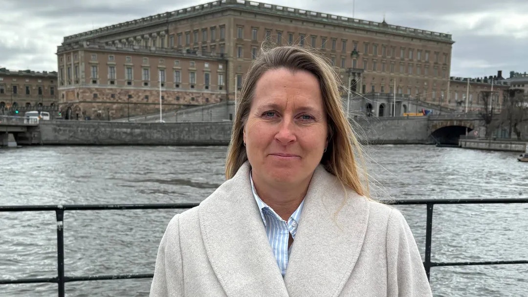 Porträtt av en kvinna i ljus kappa som står framför en vattenbild med historiska stenbyggnader i bakgrunden.