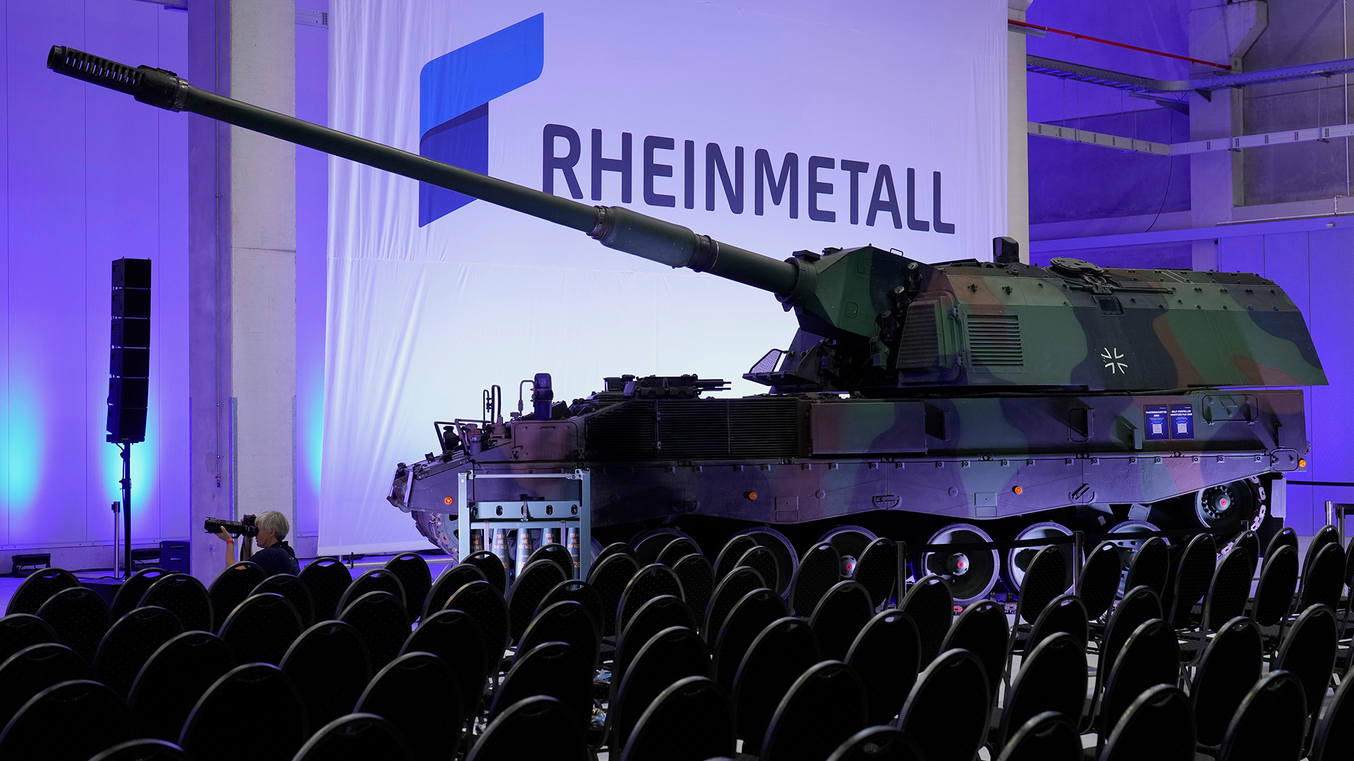 En grön militär stridsvagn från Rheinmetall utställd inomhus under blå belysning, med Rheinmetall-logon synlig i bakgrunden.