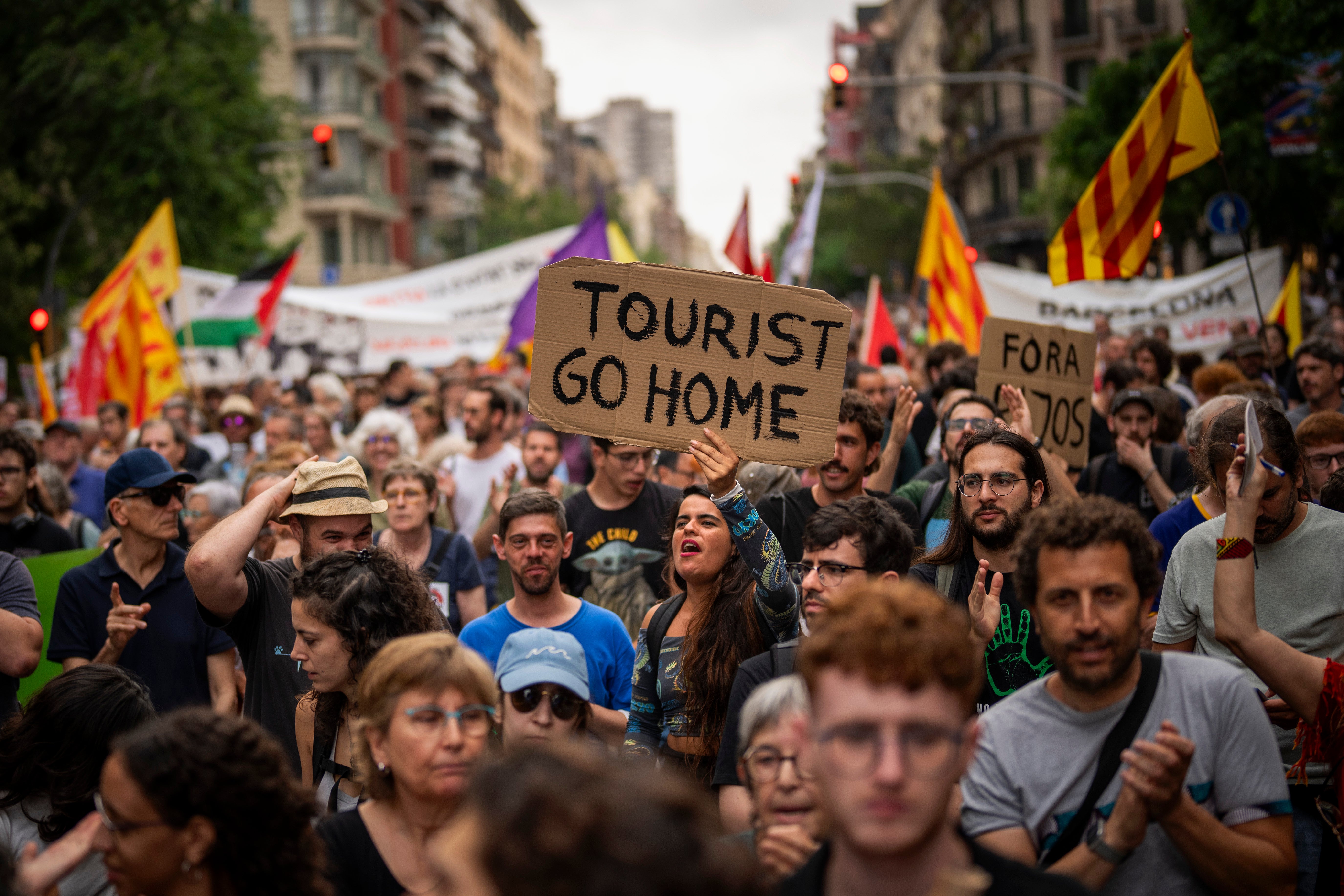 En stor demonstration i en stadsmiljö med många människor som håller upp plakat med texten "Tourist Go Home" och gula flaggor. Protesten riktar sig mot övertourism.