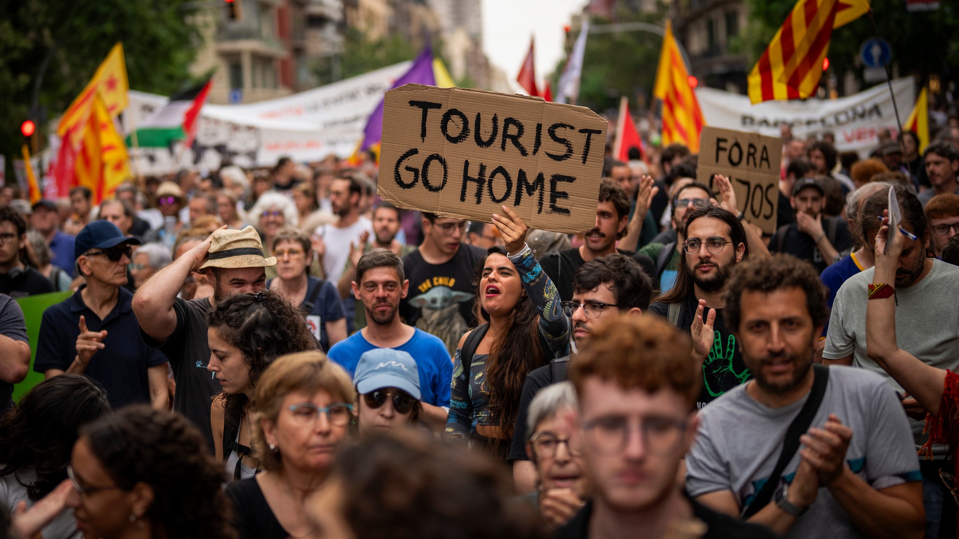 En stor demonstration i en stadsmiljö med många människor som håller upp plakat med texten "Tourist Go Home" och gula flaggor. Protesten riktar sig mot övertourism.