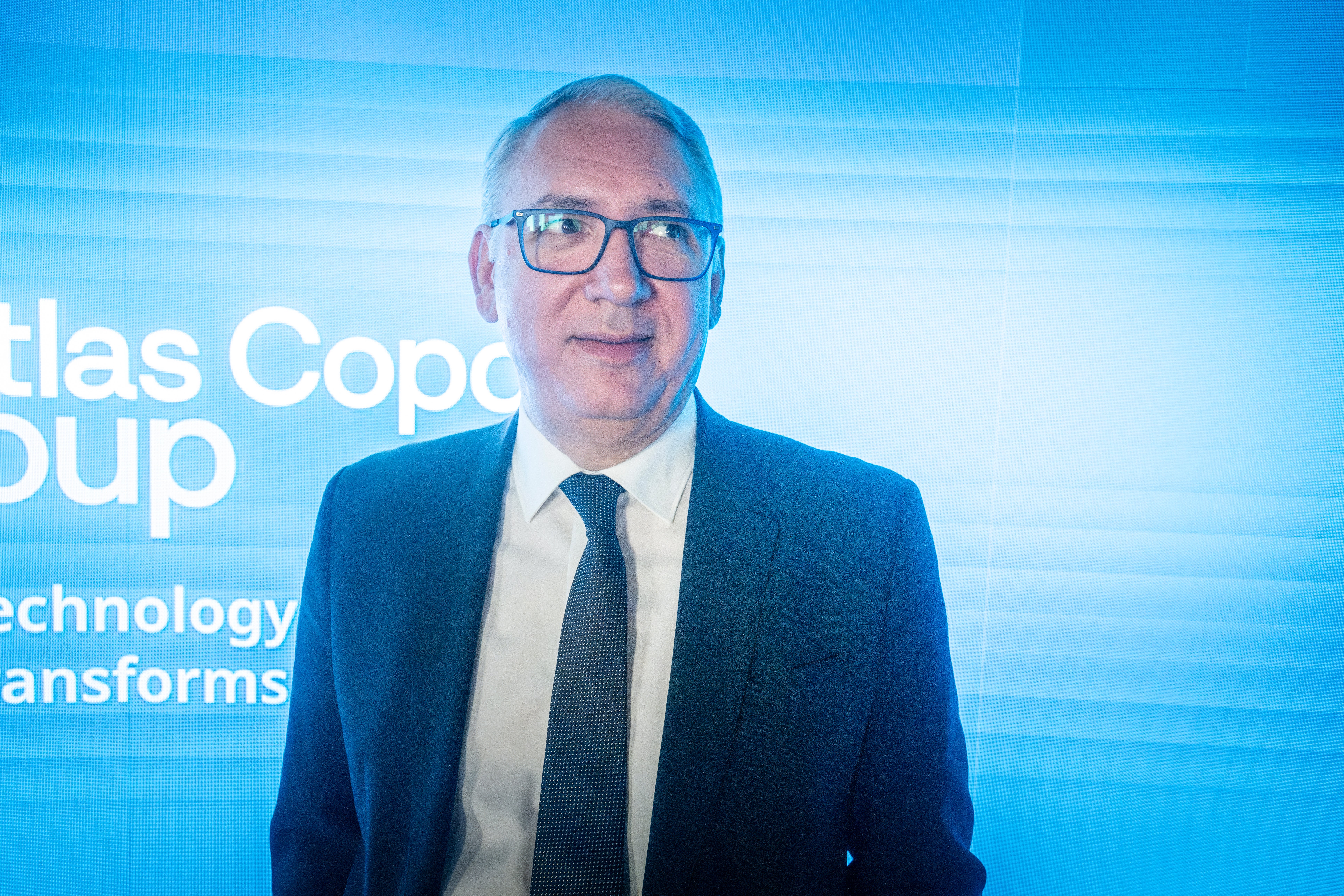 En man i glasögon och blå kostym framför en blå bakgrund med text.