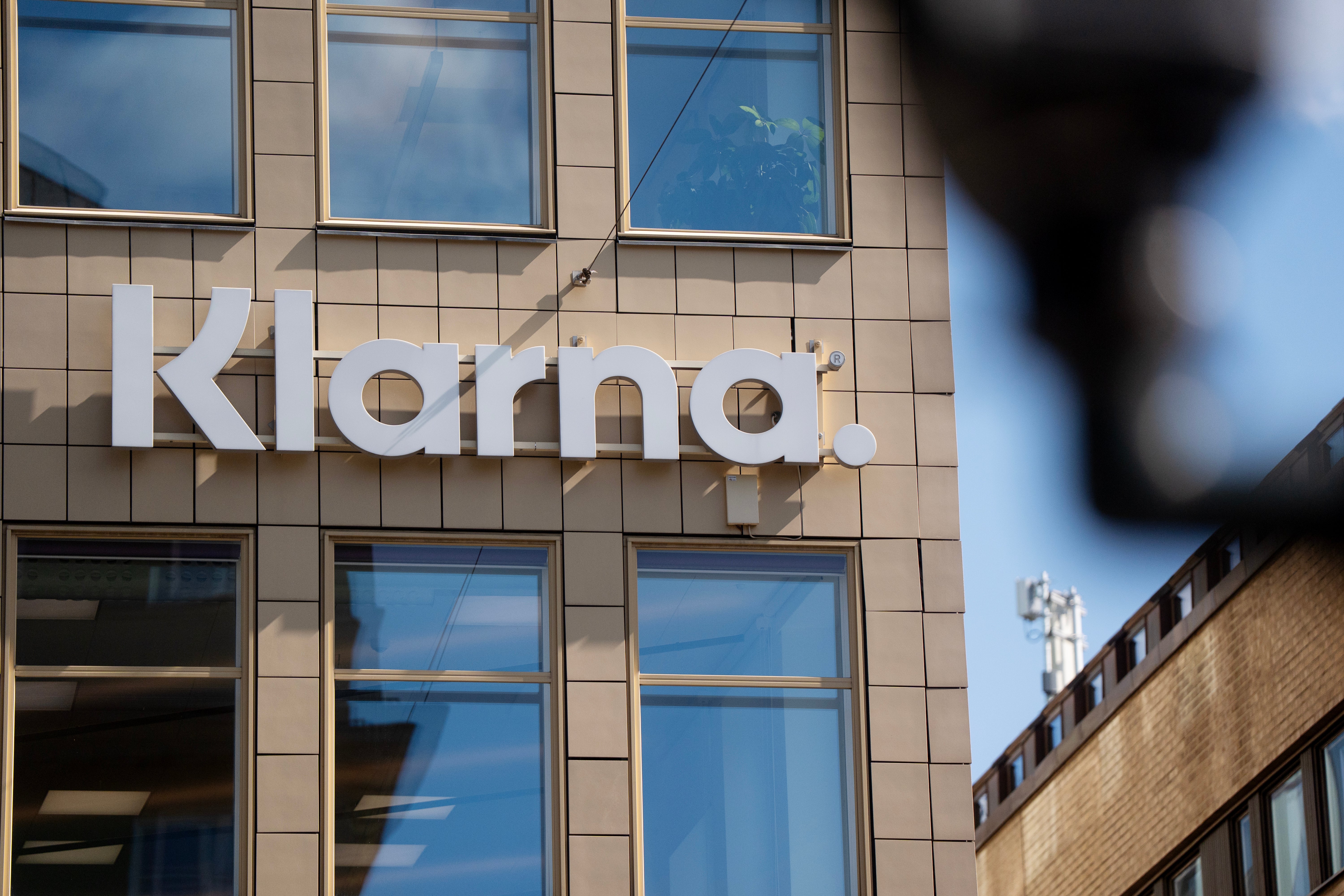 Vita bokstäver med Klarna-logotypen på en beige byggnadsfasad med stora fönster.