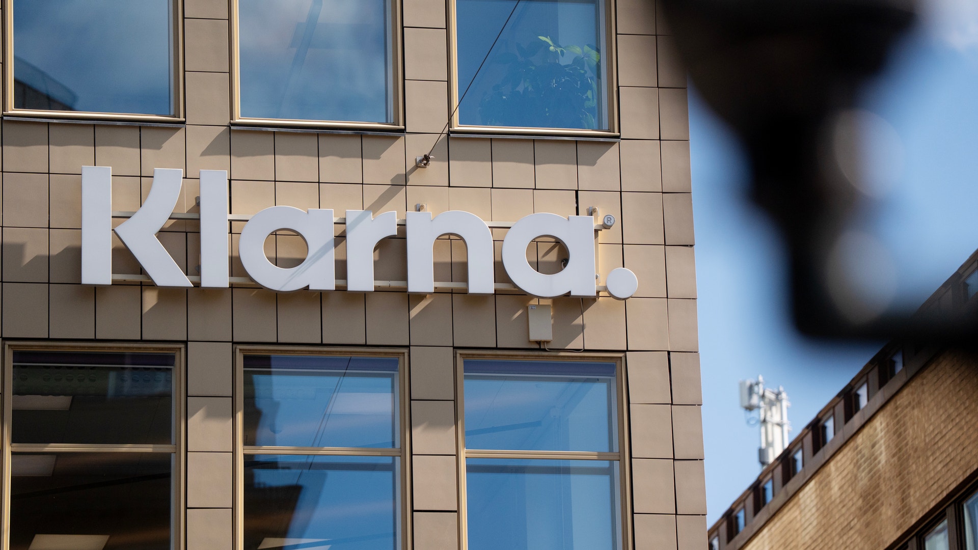 Vita bokstäver med Klarna-logotypen på en beige byggnadsfasad med stora fönster.