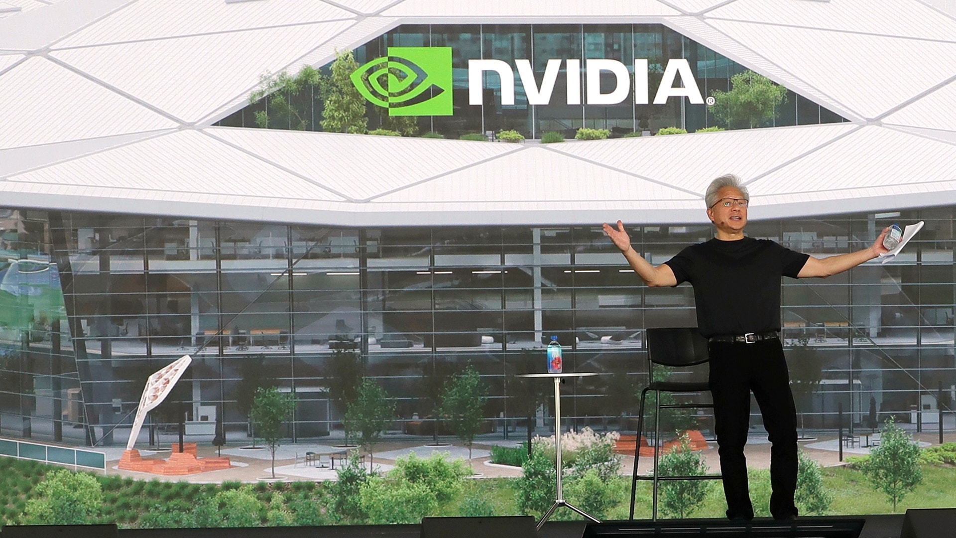 Man i svart kläder står framför NVIDIAs huvudkontor med armarna utsträckta. NVIDIAs gröna logotyp är synlig på taket ovanför glasfasaden.
