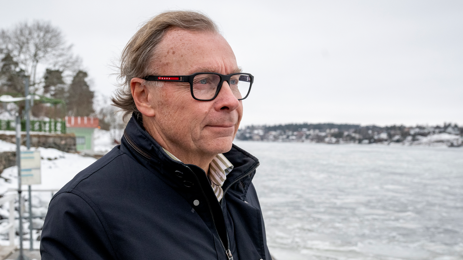Porträttfoto av Peter Malmqvist, en man med glasögon och mörk jacka, fotograferad vid en vattenbrygg med träd och bostäder synliga i bakgrunden.