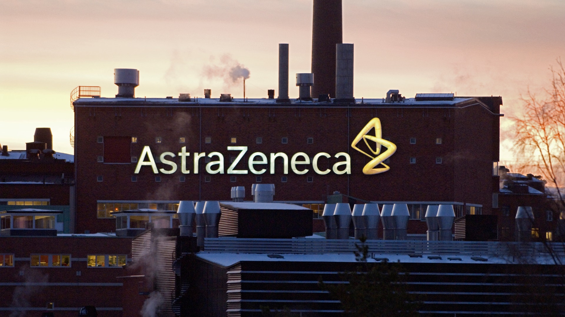Industribyggnad med AstraZenecas logo och namn belyst på fasaden vid skymning.