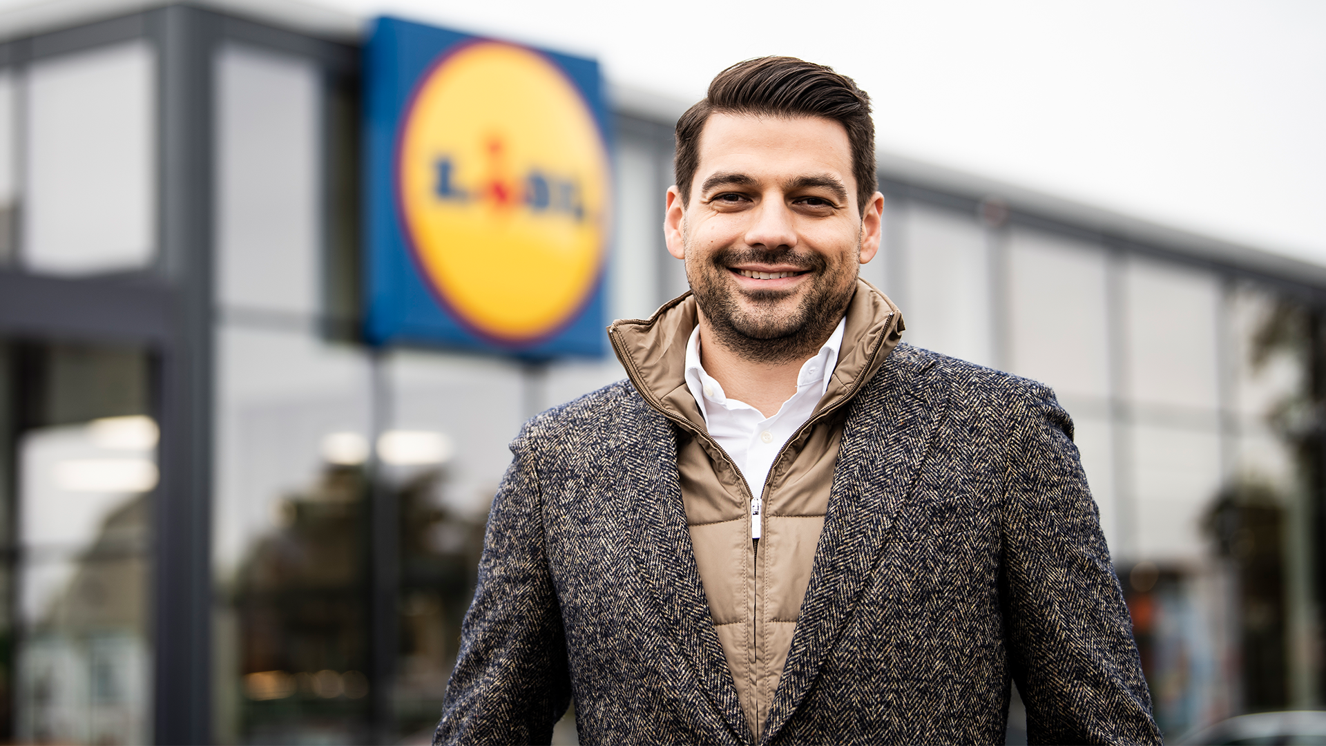 Porträtt av en leende man i grå jacka som står framför en Lidl-butik.