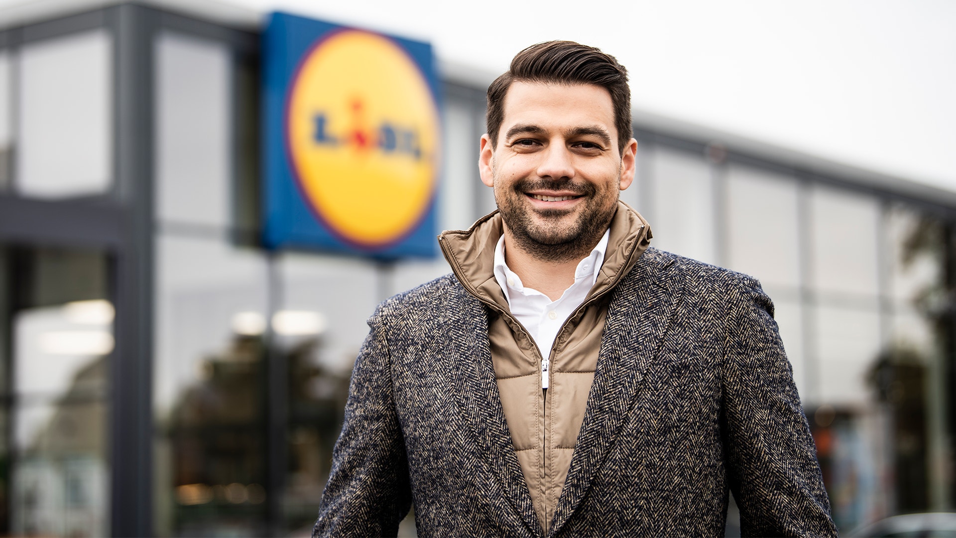Porträtt av en leende man i grå jacka som står framför en Lidl-butik.