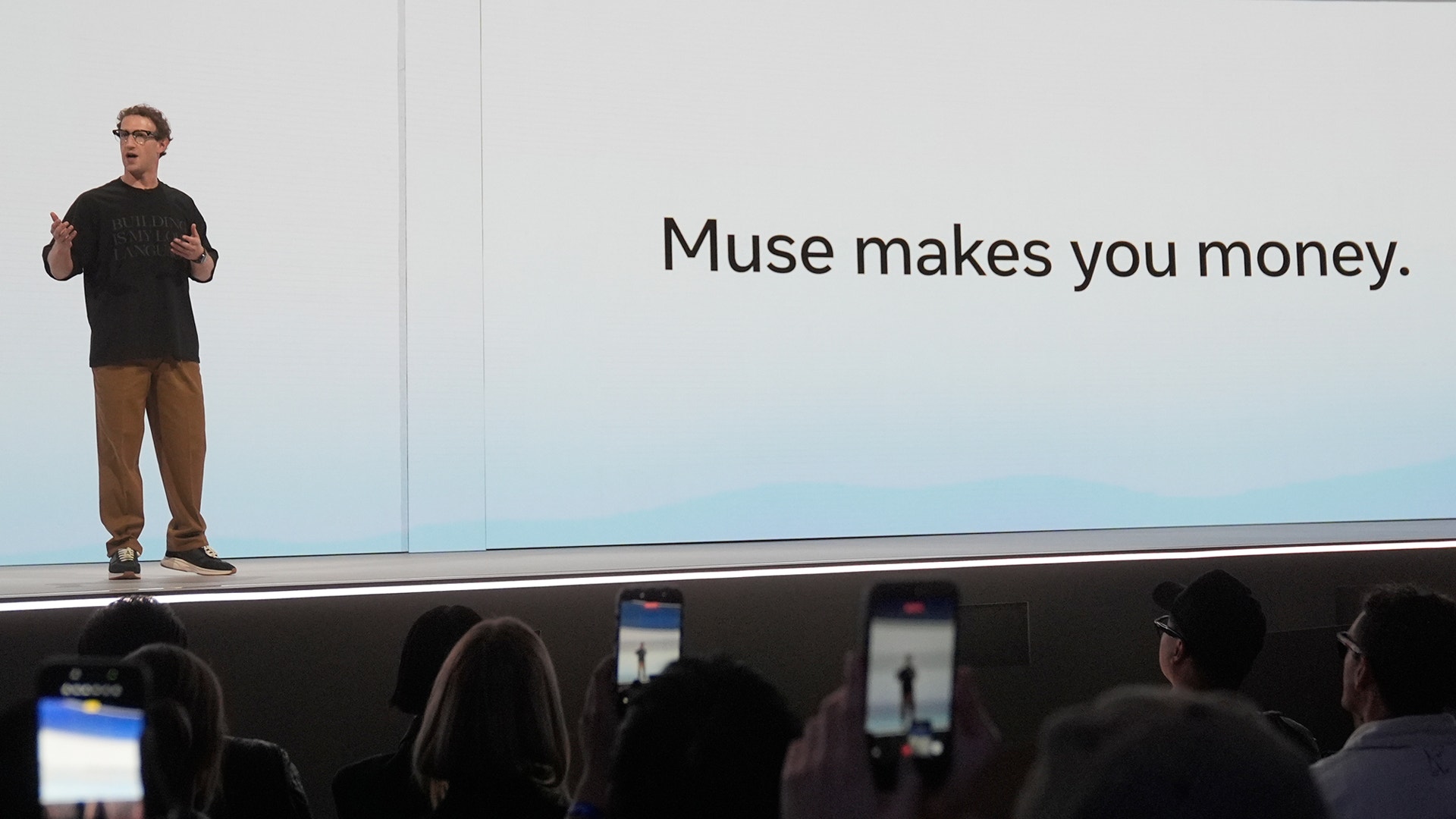 En man håller en presentation på en scen inför publik, med texten "Muse makes you money" projicerad bakom sig.