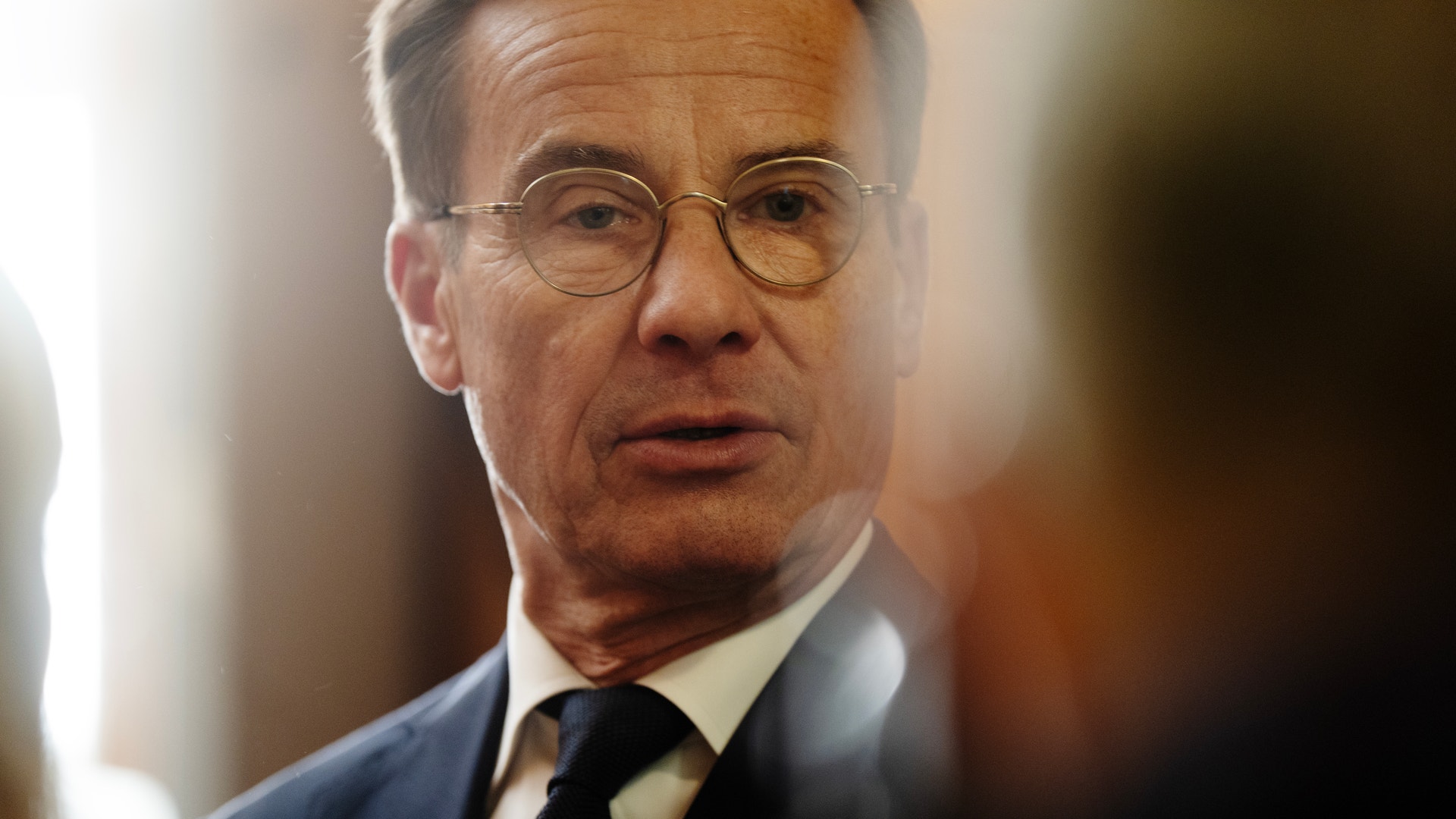 Man med glasögon och kostym pratar med människor omkring sig.