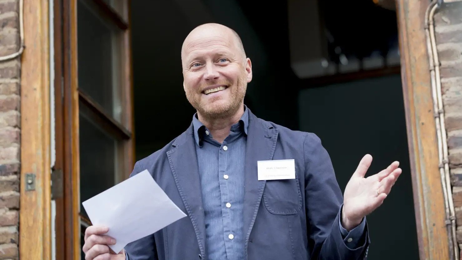 En man i mörkblå skjorta står i en dörröppning och ler. Han håller ett papper i ena handen och gestikulerar välkomnande med den andra.