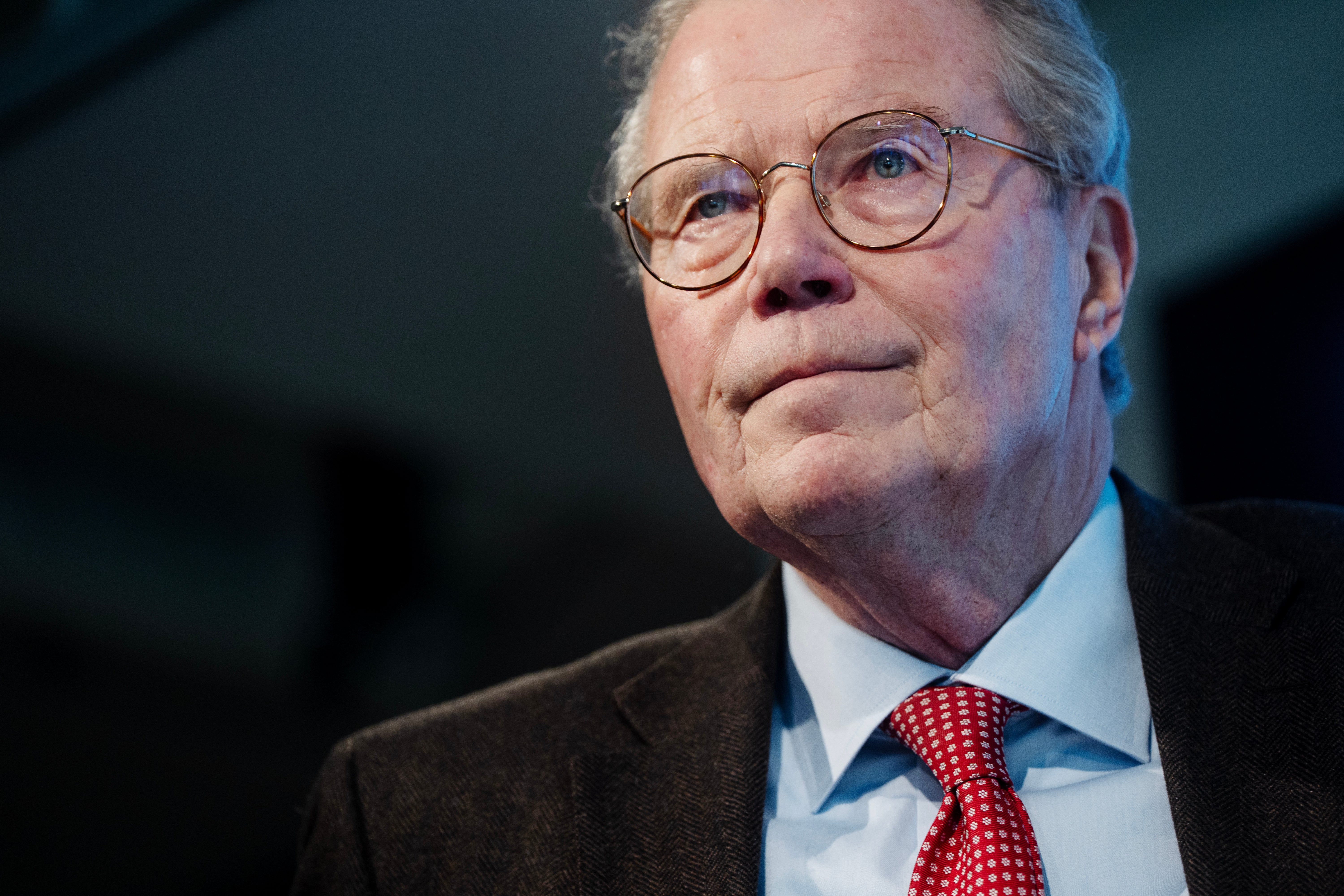 Porträtt av en äldre man med glasögon, mörkblå kostym, ljusblå skjorta och röd slips mot mörk bakgrund.