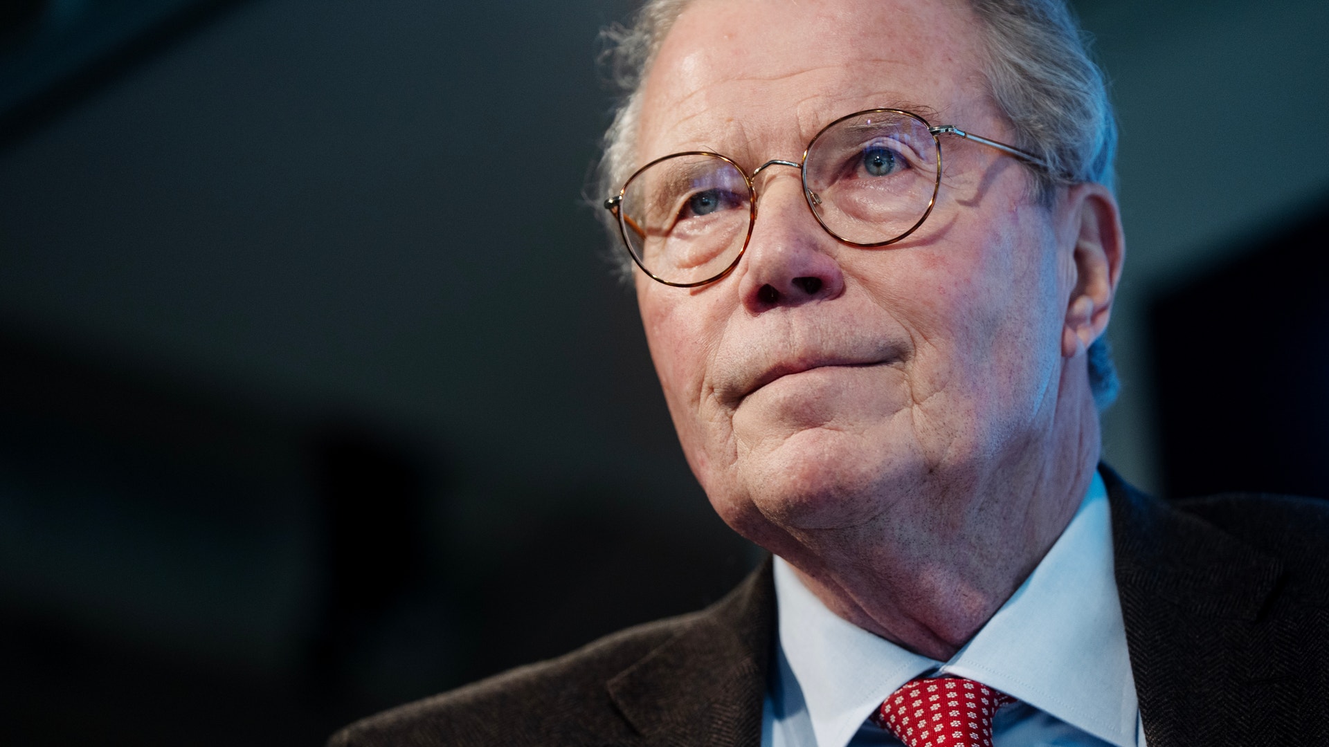 Porträtt av en äldre man med glasögon, mörkblå kostym, ljusblå skjorta och röd slips mot mörk bakgrund.