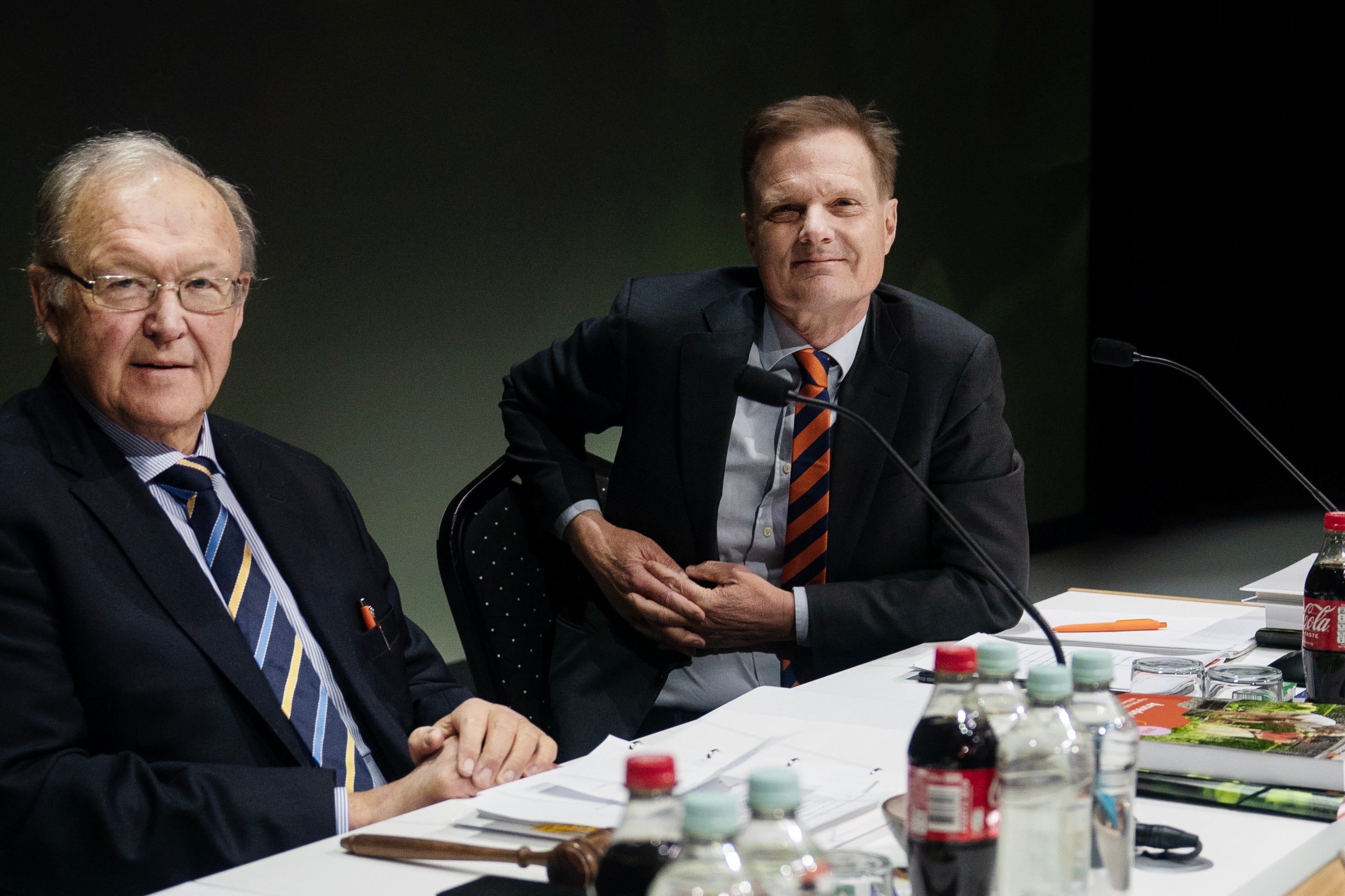 Två män i affärskläder sitter vid ett bord under en professionell möte eller paneldiskussion.