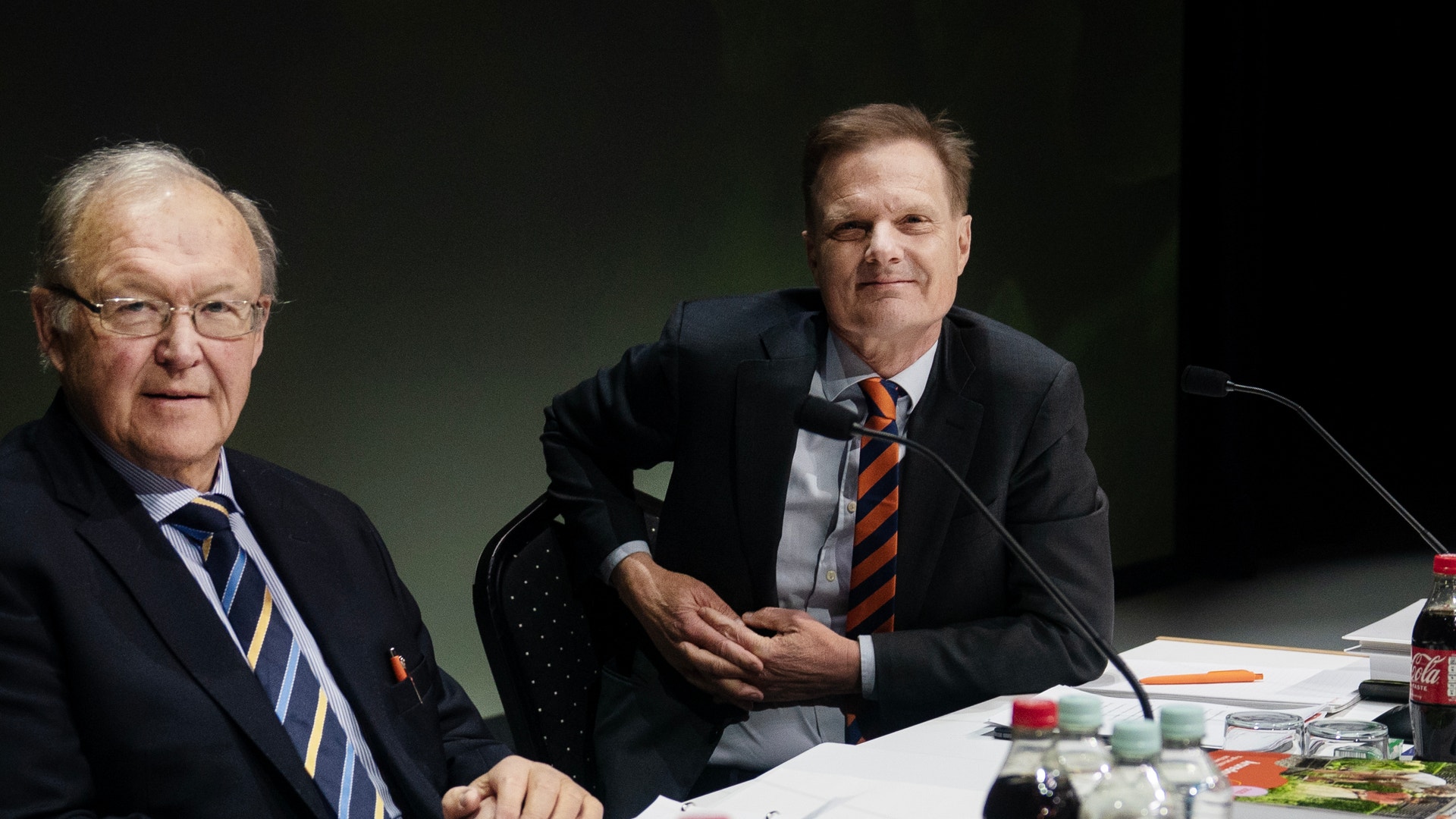 Två män i affärskläder sitter vid ett bord under en professionell möte eller paneldiskussion.