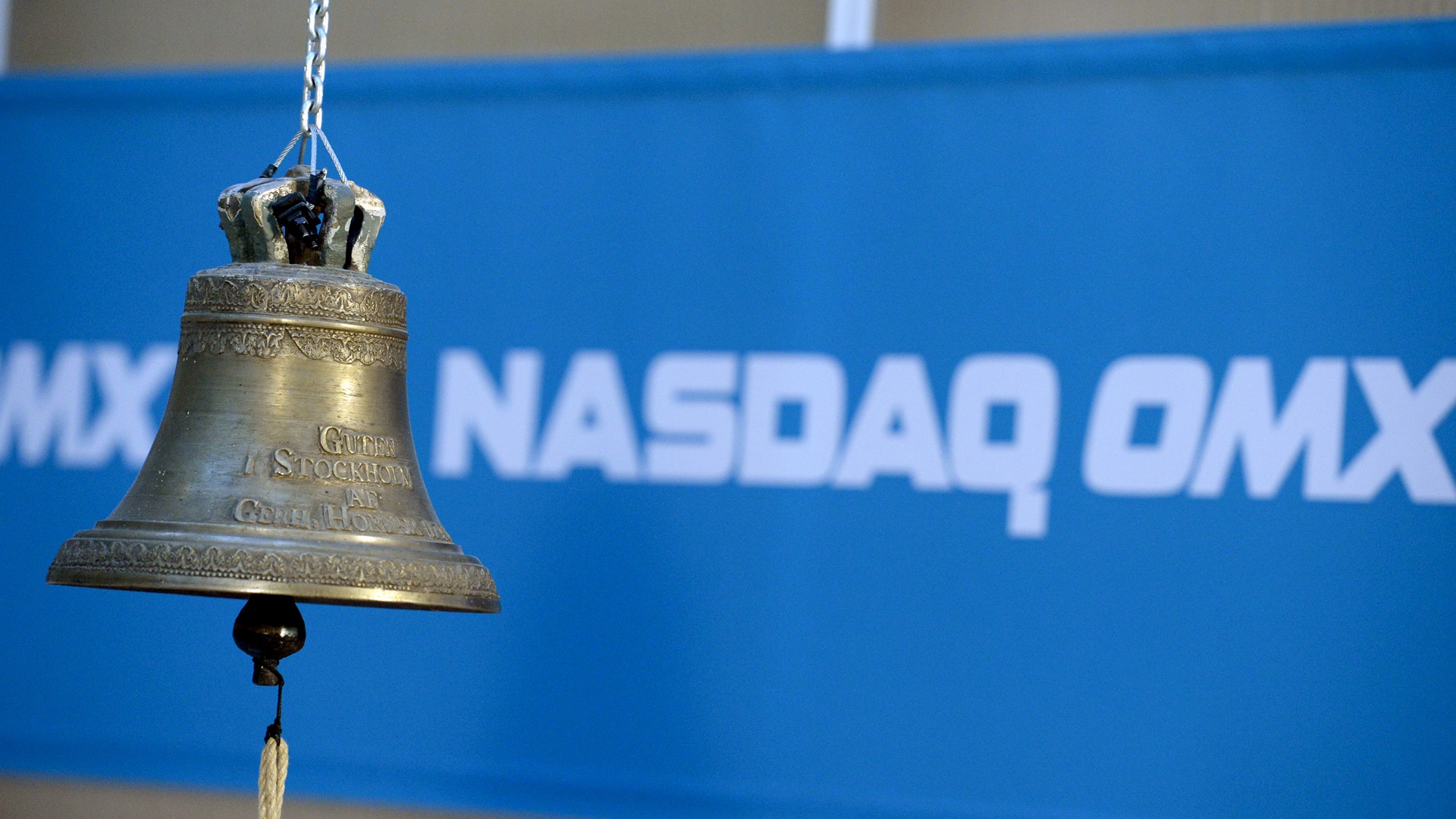 En mässingsklocka hängande framför en blå vägg med NASDAQ OMX-logotyp.