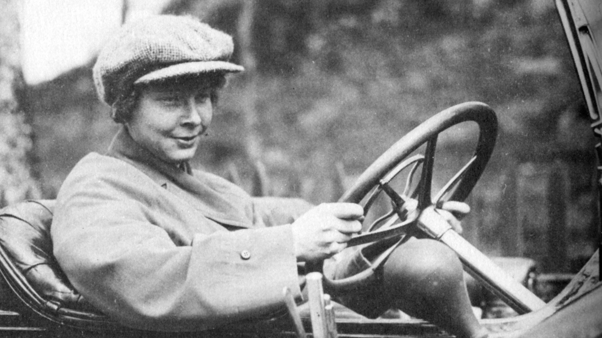 Svartvit historisk fotografi av en ung person som sitter i en öppen bil från början av 1900-talet och håller i ratten. Personen bär en flat cap och ser glad ut.