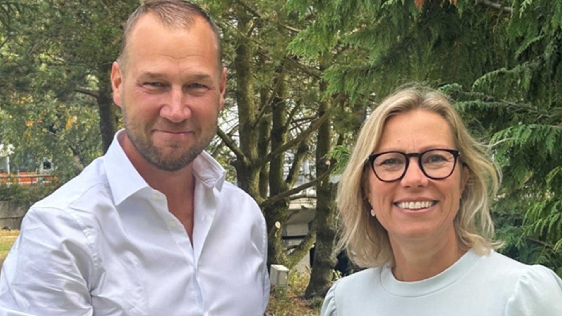 Två personer som står tillsammans och ler mot kameran. En man i vit skjorta och en kvinna i ljus tröja och glasögon. Gröna träd syns i bakgrunden.