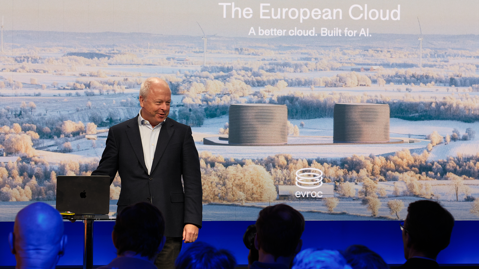 En man i kostym presenterar på en scen framför en skärm som visar ett flygfotografi av ett snöigt landskap med industriella byggnader.