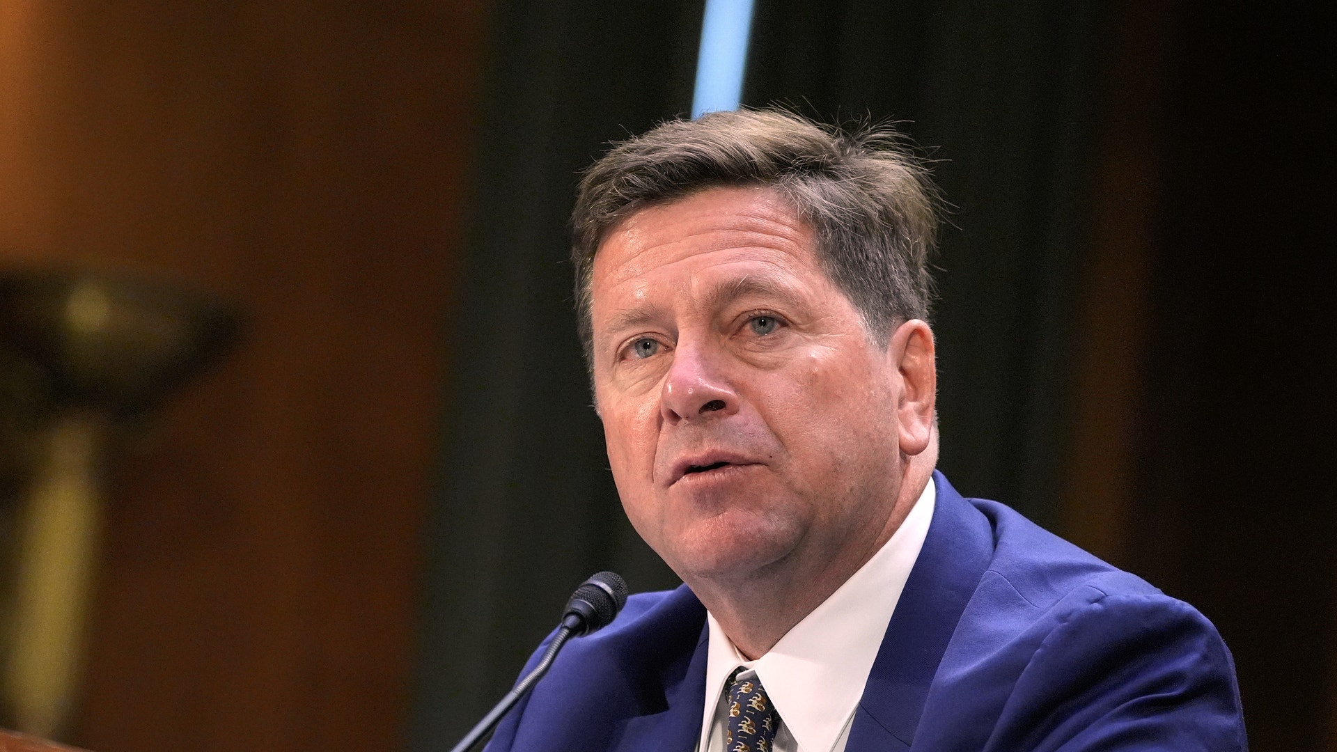 En man i kostym talar i en mikrofon under en utfrågning.