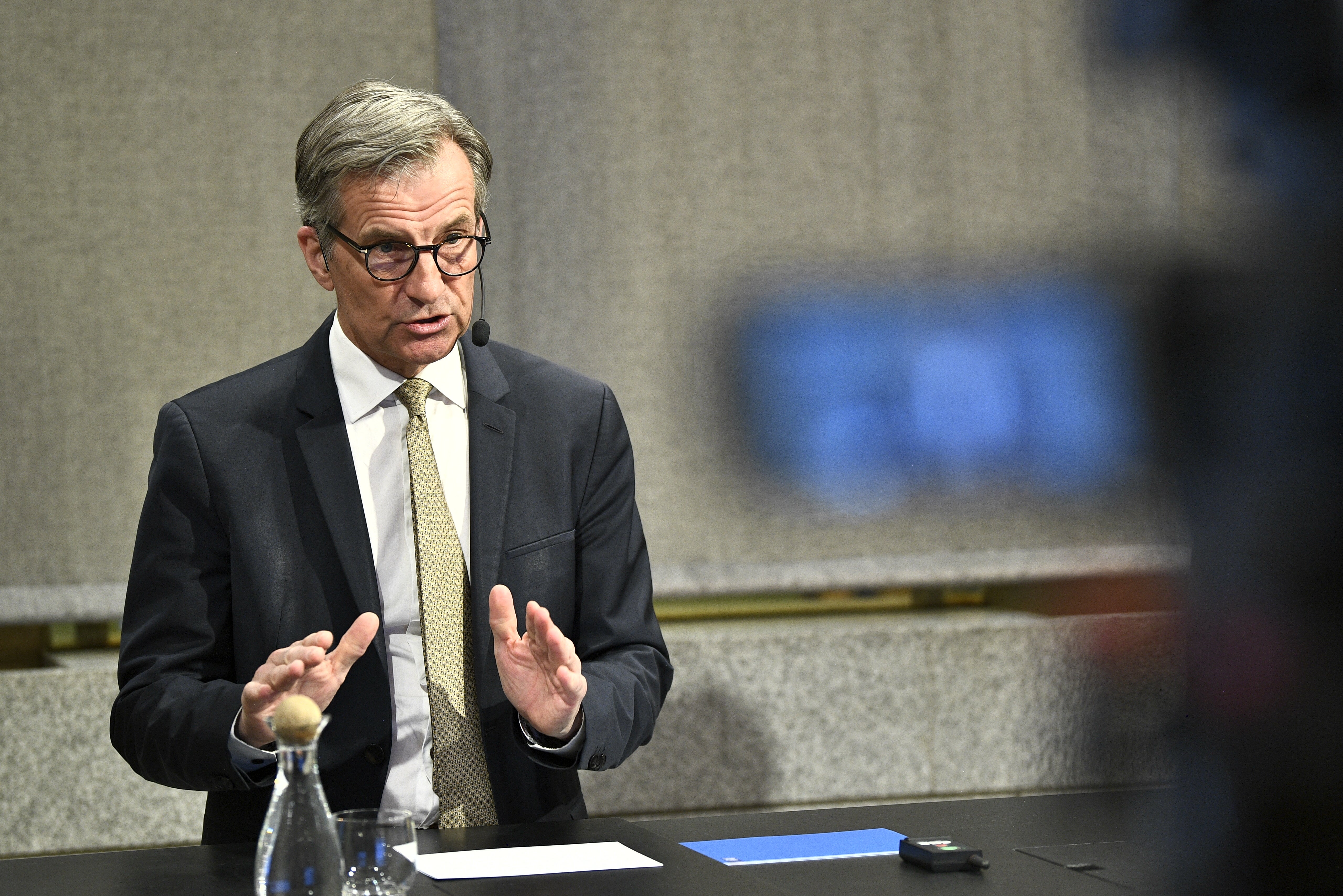 En man i kostym och glasögon talar vid ett möte, med händerna upphöjda i en gestikulerande rörelse.