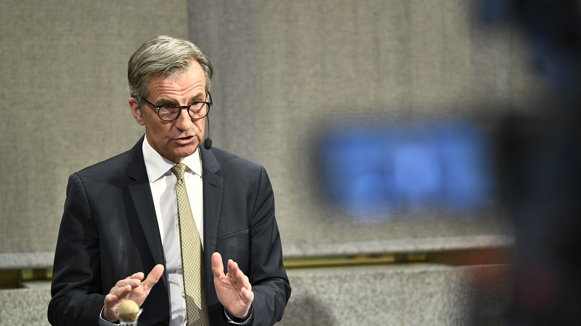 En man i kostym och glasögon talar vid ett möte, med händerna upphöjda i en gestikulerande rörelse.