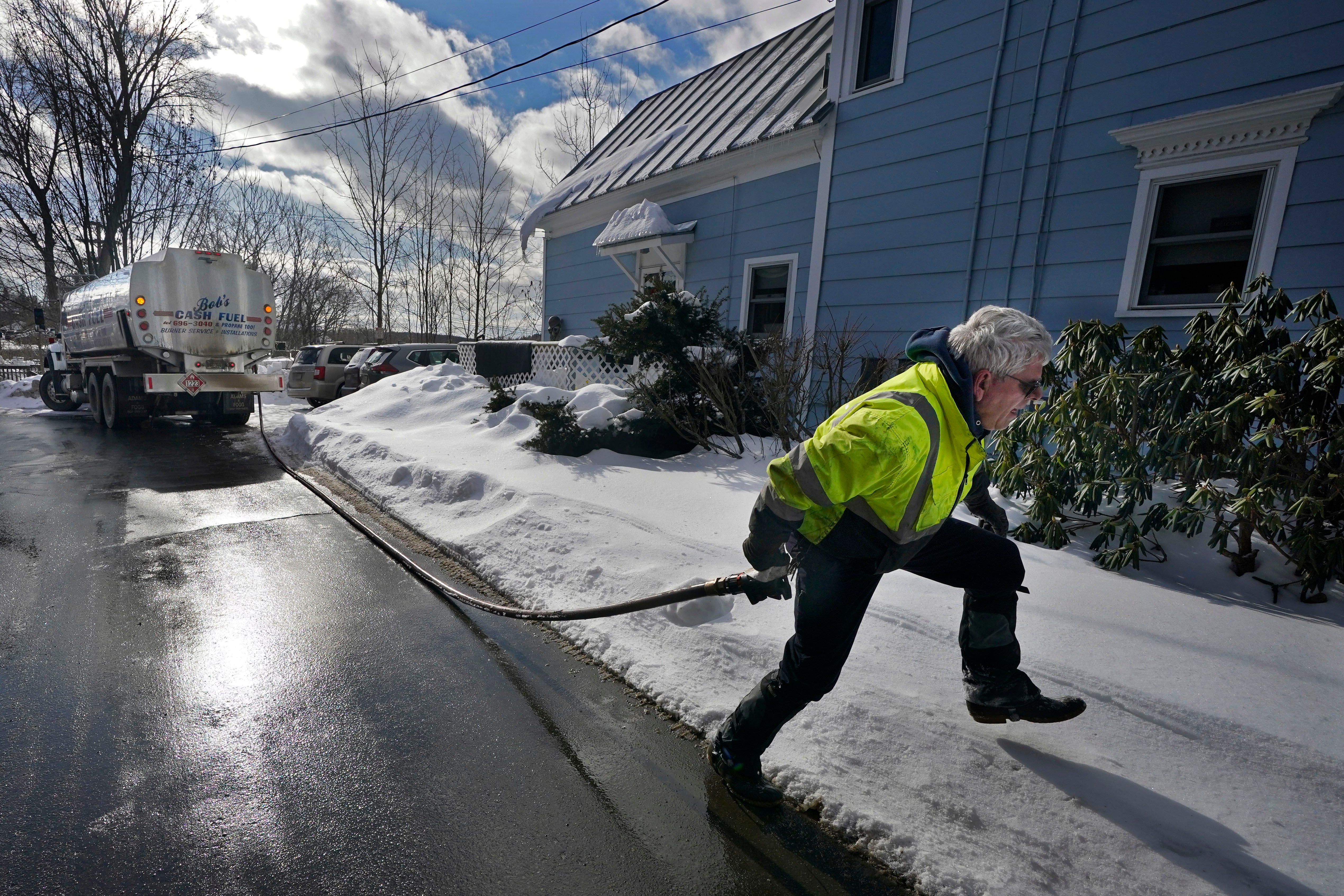 En man i gul reflexjacka drar en slang från en tankbil längs en snöig villagata, framför ett blått hus.