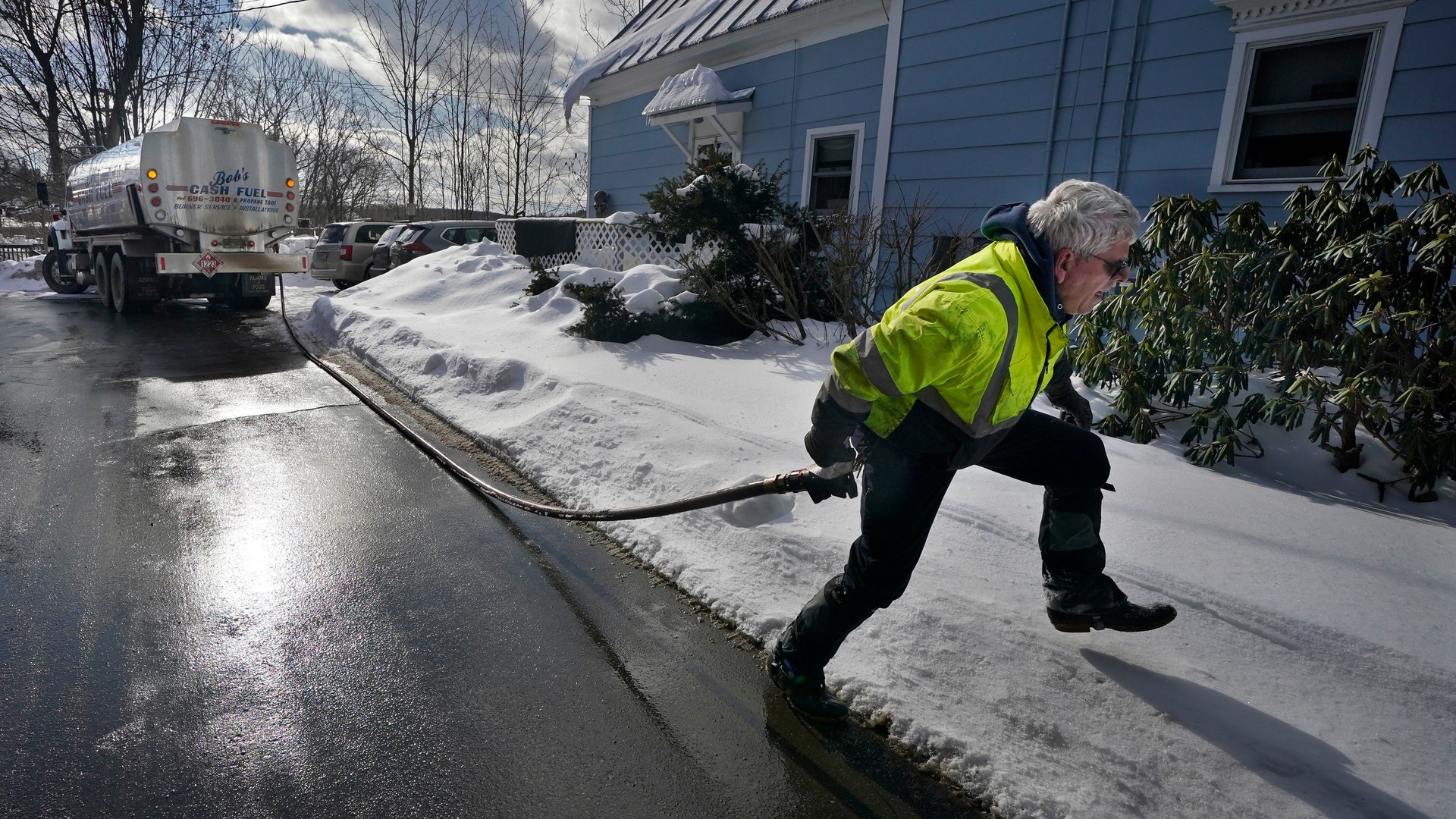 En man i gul reflexjacka drar en slang från en tankbil längs en snöig villagata, framför ett blått hus.