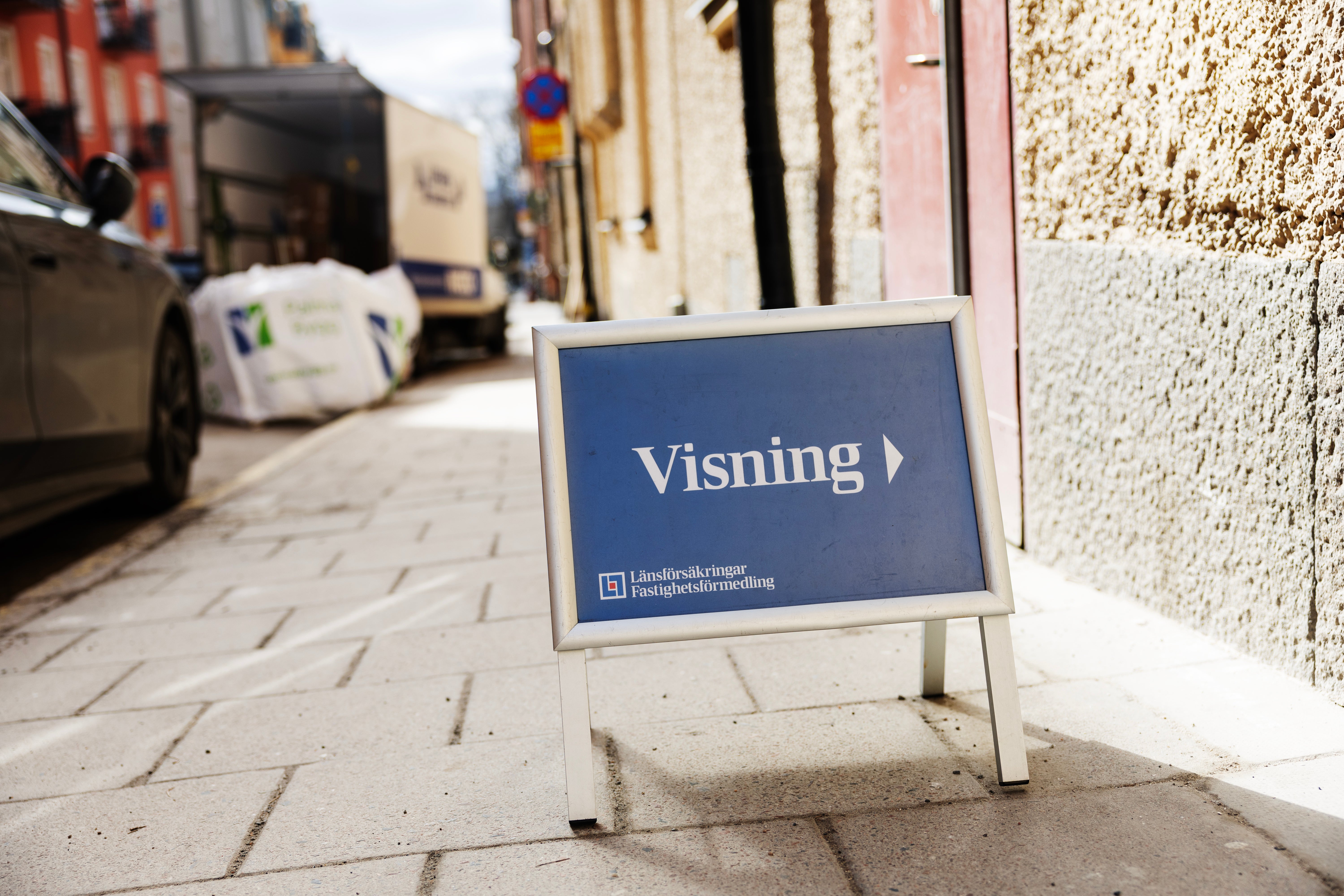 En blå skylt med texten 'Visning' står på en gata framför ett hus. Skyltens logotyp indikerar att det är en fastighetsvisning.