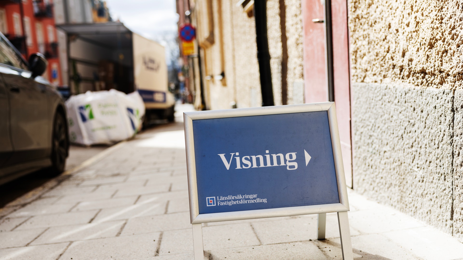 En blå skylt med texten 'Visning' står på en gata framför ett hus. Skyltens logotyp indikerar att det är en fastighetsvisning.