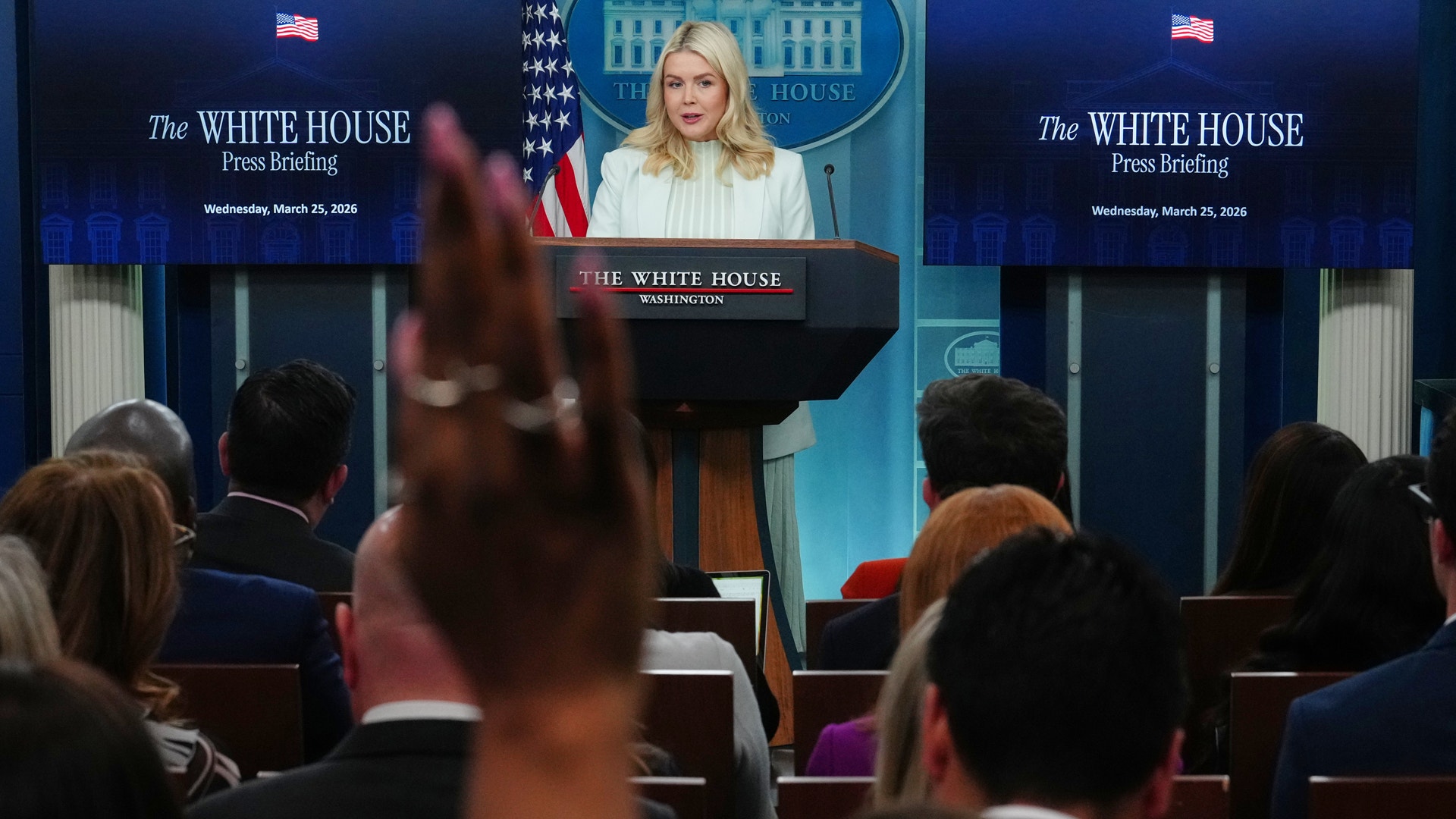 En kvinna står vid ett podium under en presskonferens på Vita huset, omgiven av journalister och amerikanska flaggor.