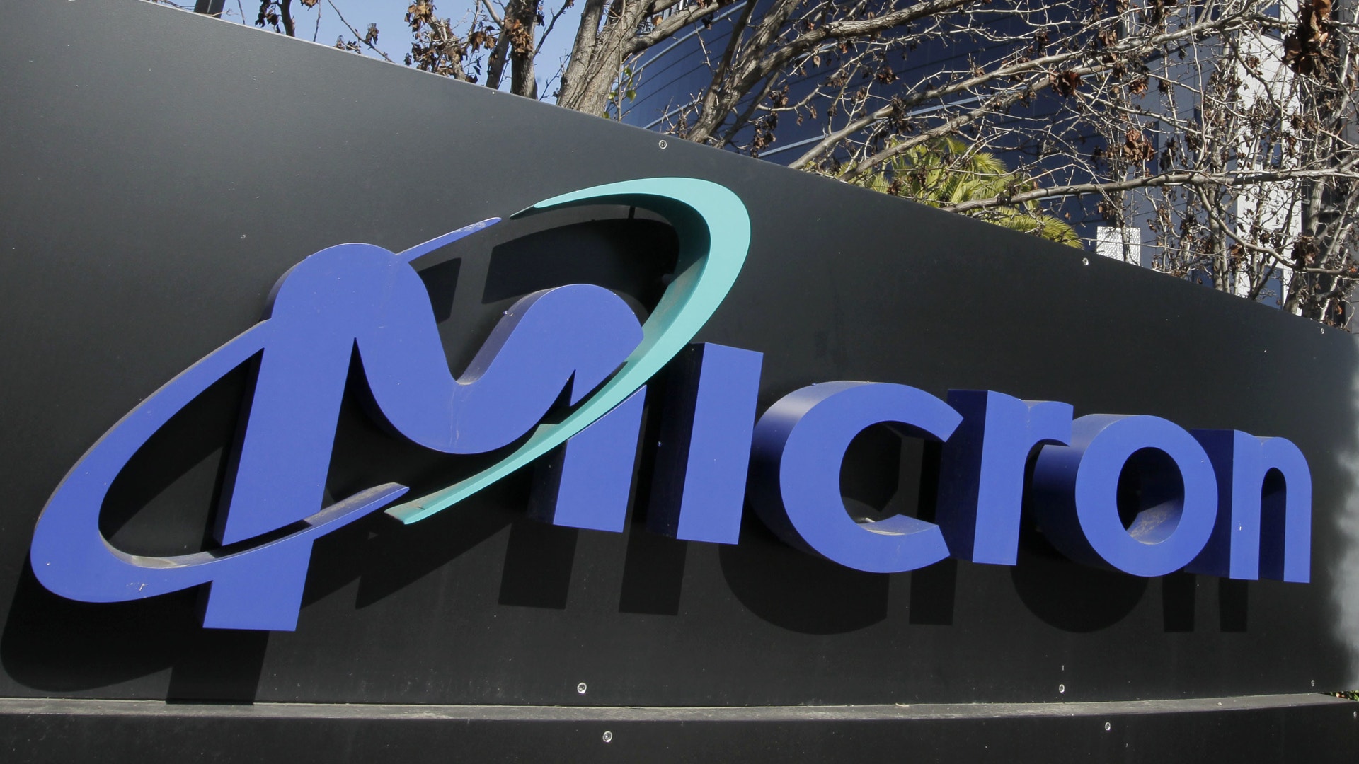 Företagsskylt med Microns logotyp i blå bokstäver och grön båge på svart bakgrund