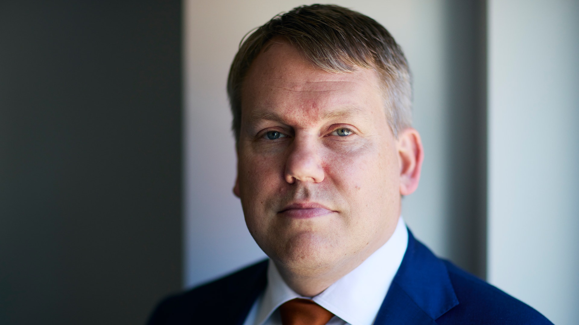 Porträtt av en man i mörk kostym och brun slips mot en suddig bakgrund.