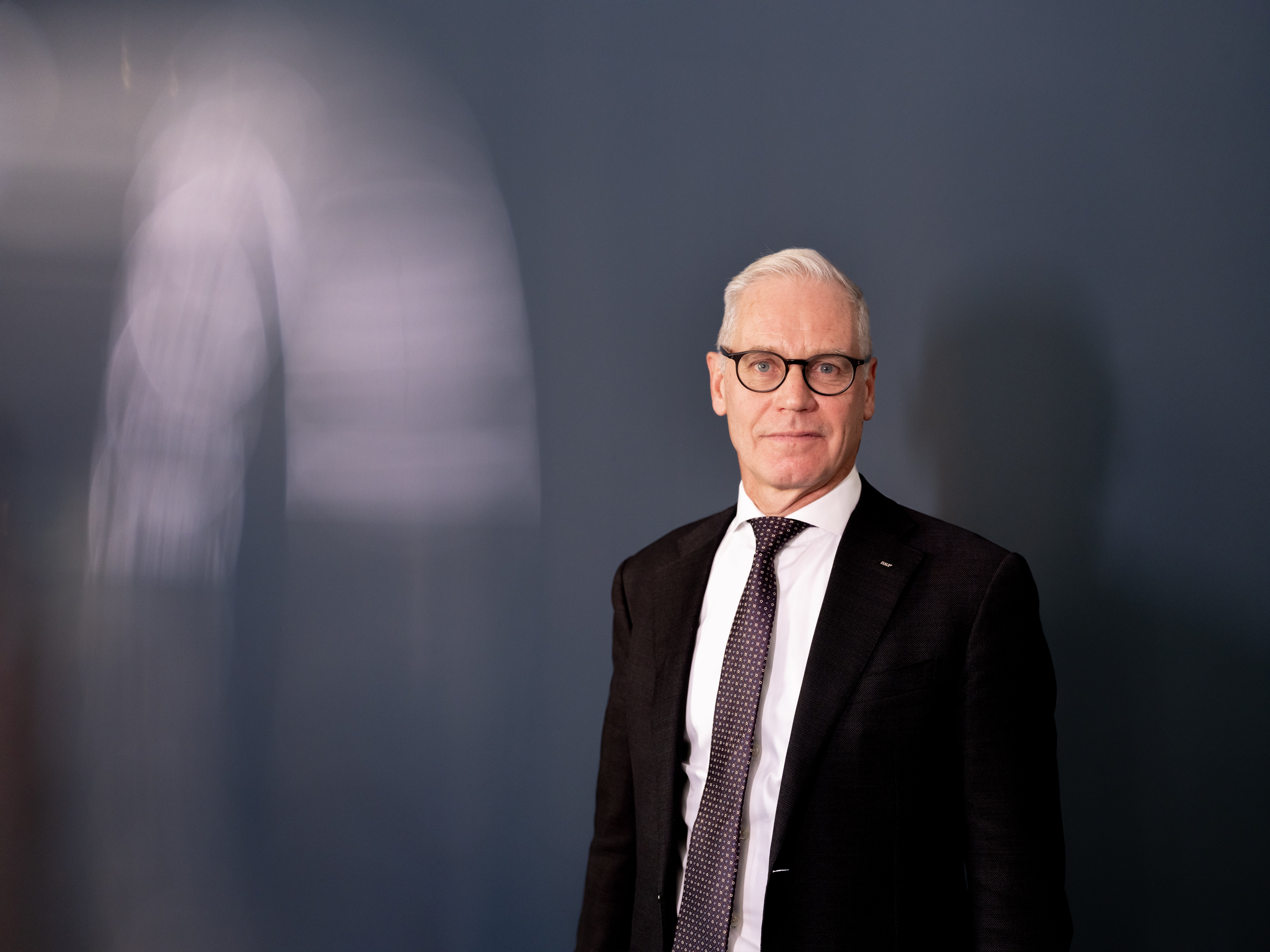 En äldre man i kostym och slips står framför en mörk vägg.