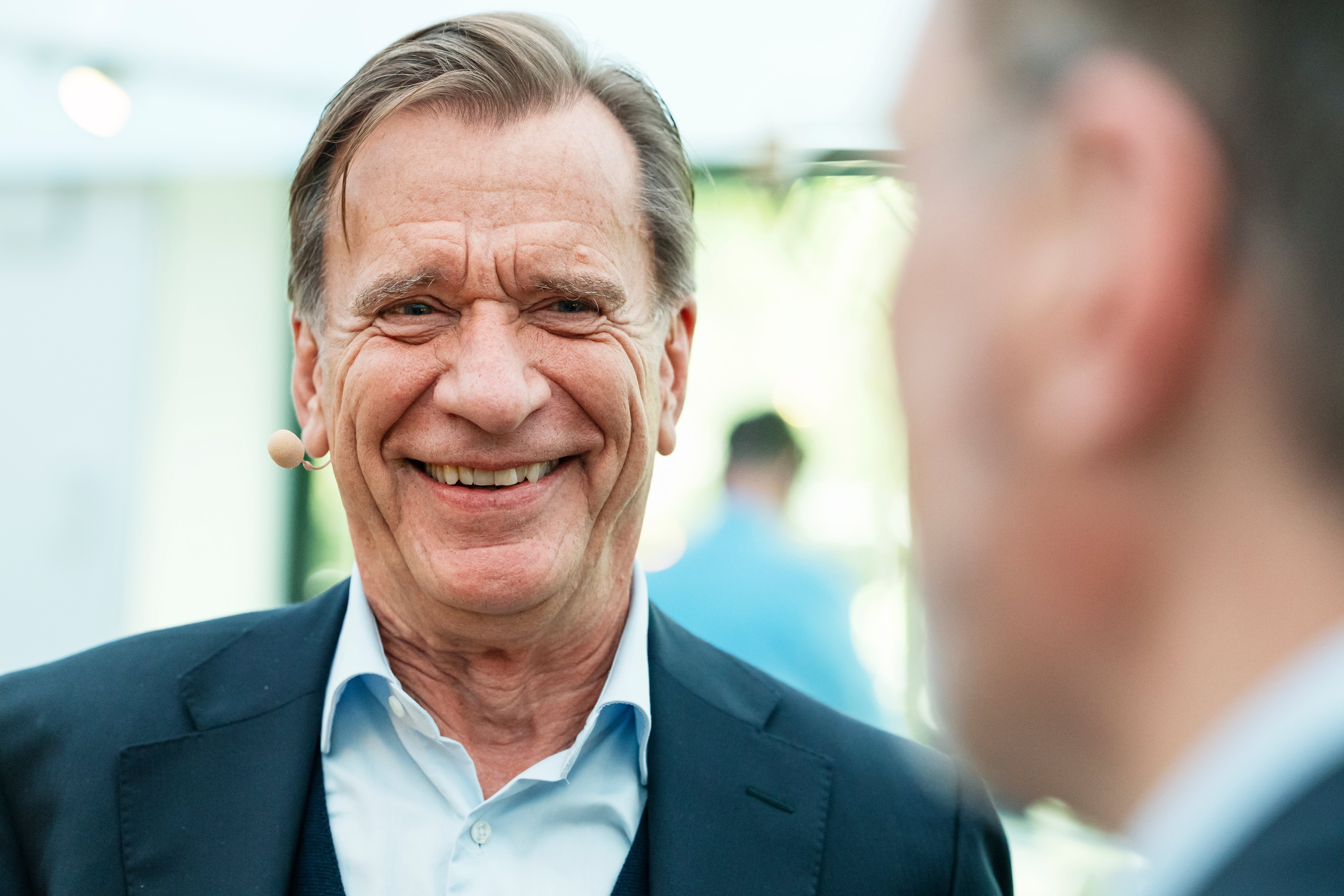 En leende äldre man i kostym med mikrofon, i samtal med en person utomhus.