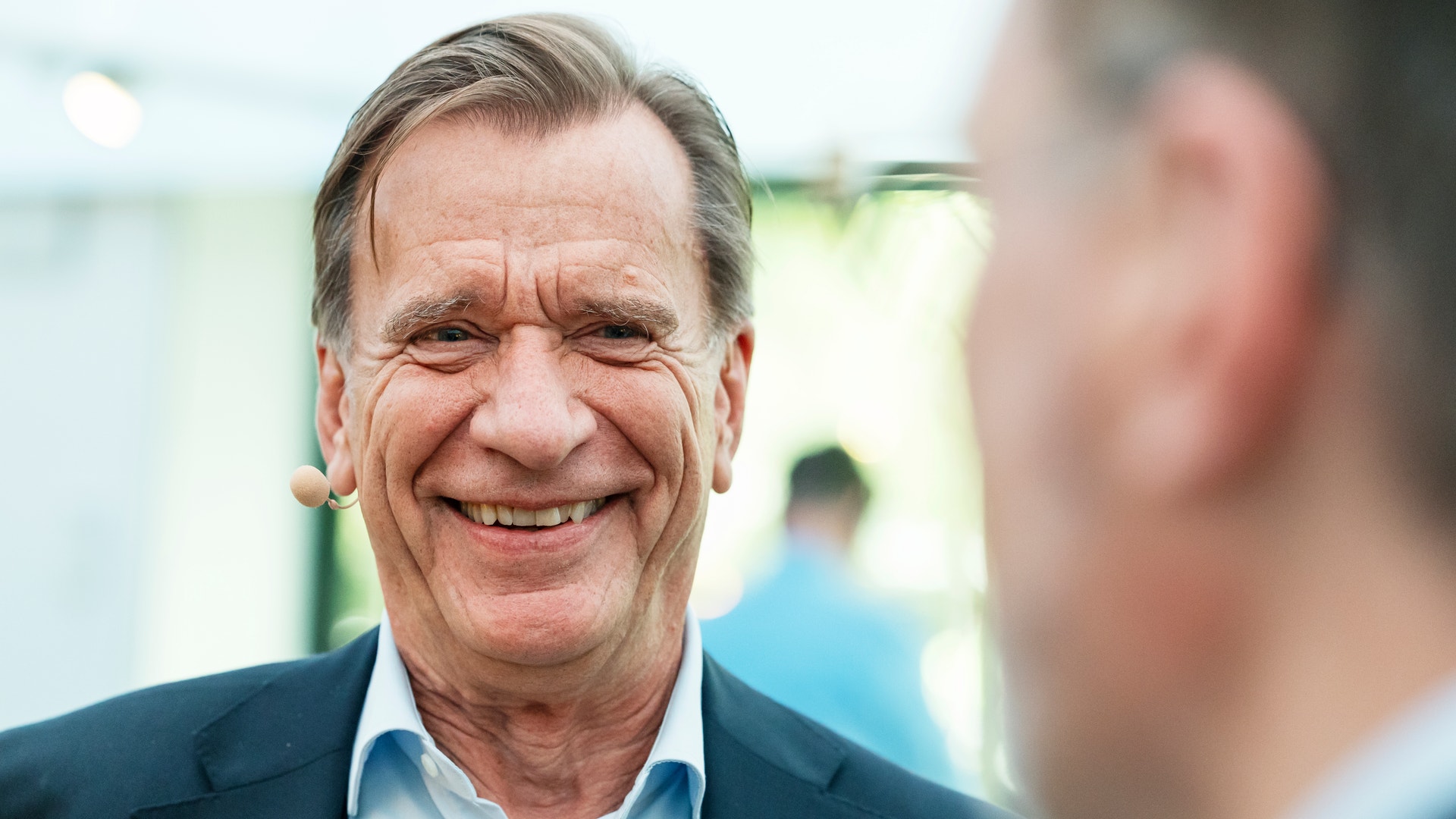 En leende äldre man i kostym med mikrofon, i samtal med en person utomhus.