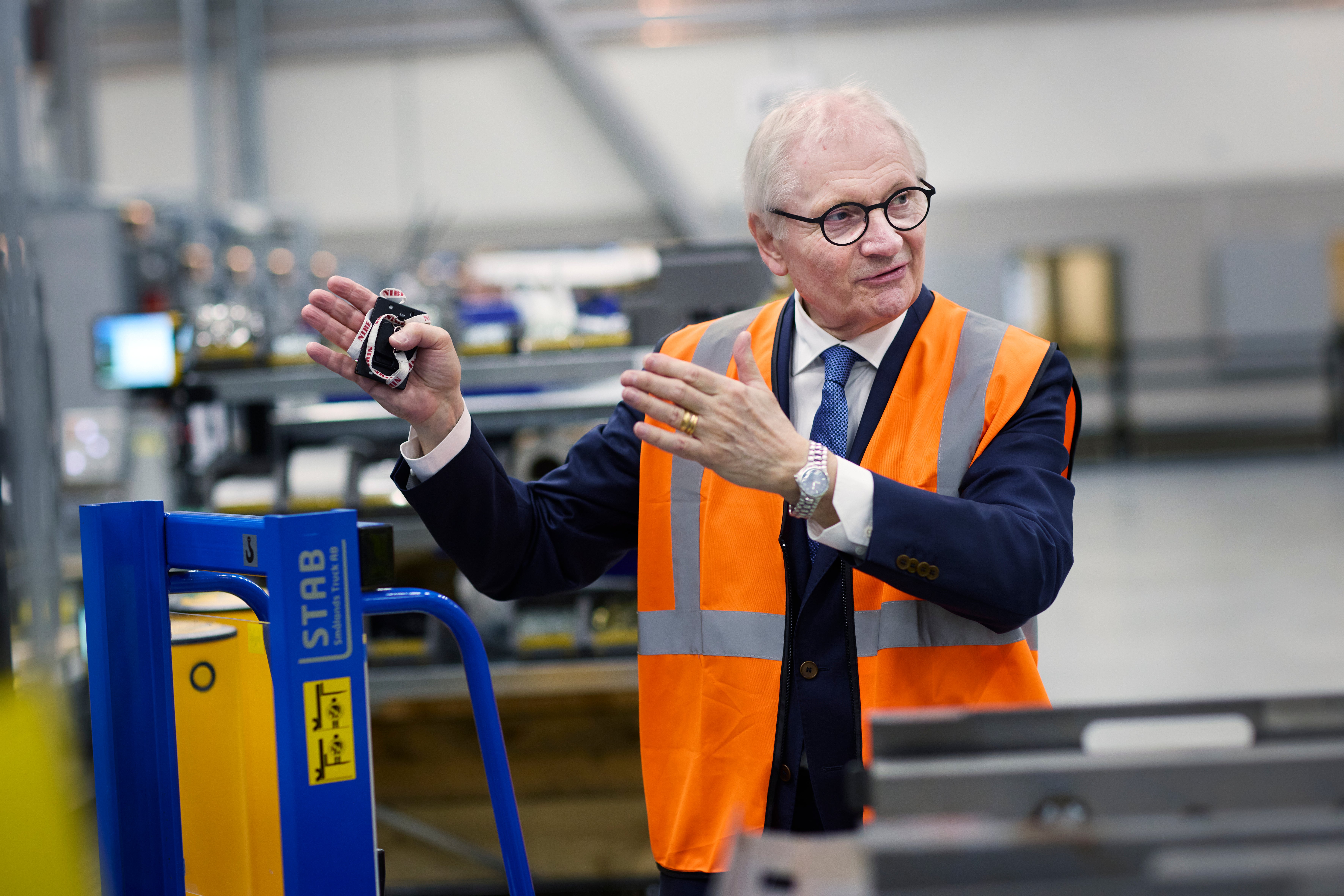 En äldre man i säkerhetväst och kostym står i en fabrik och presenterar en maskinkomponent.