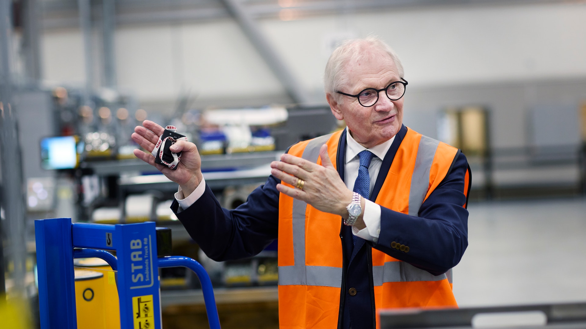 En äldre man i säkerhetväst och kostym står i en fabrik och presenterar en maskinkomponent.